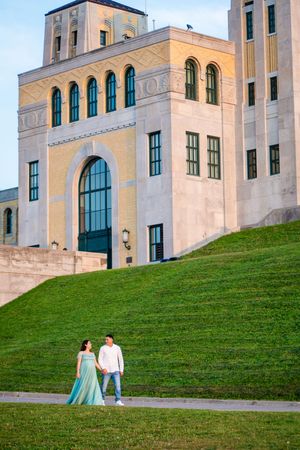 Couple holding hands in front of a grand historic building with arched windows and green lawn at golden hour. toronto