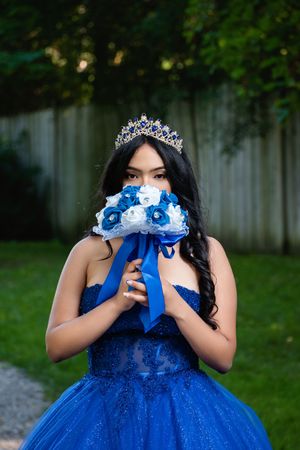 Young woman in blue quinceañera gown and crown holds blue and white bouquet covering her face outdoors.
