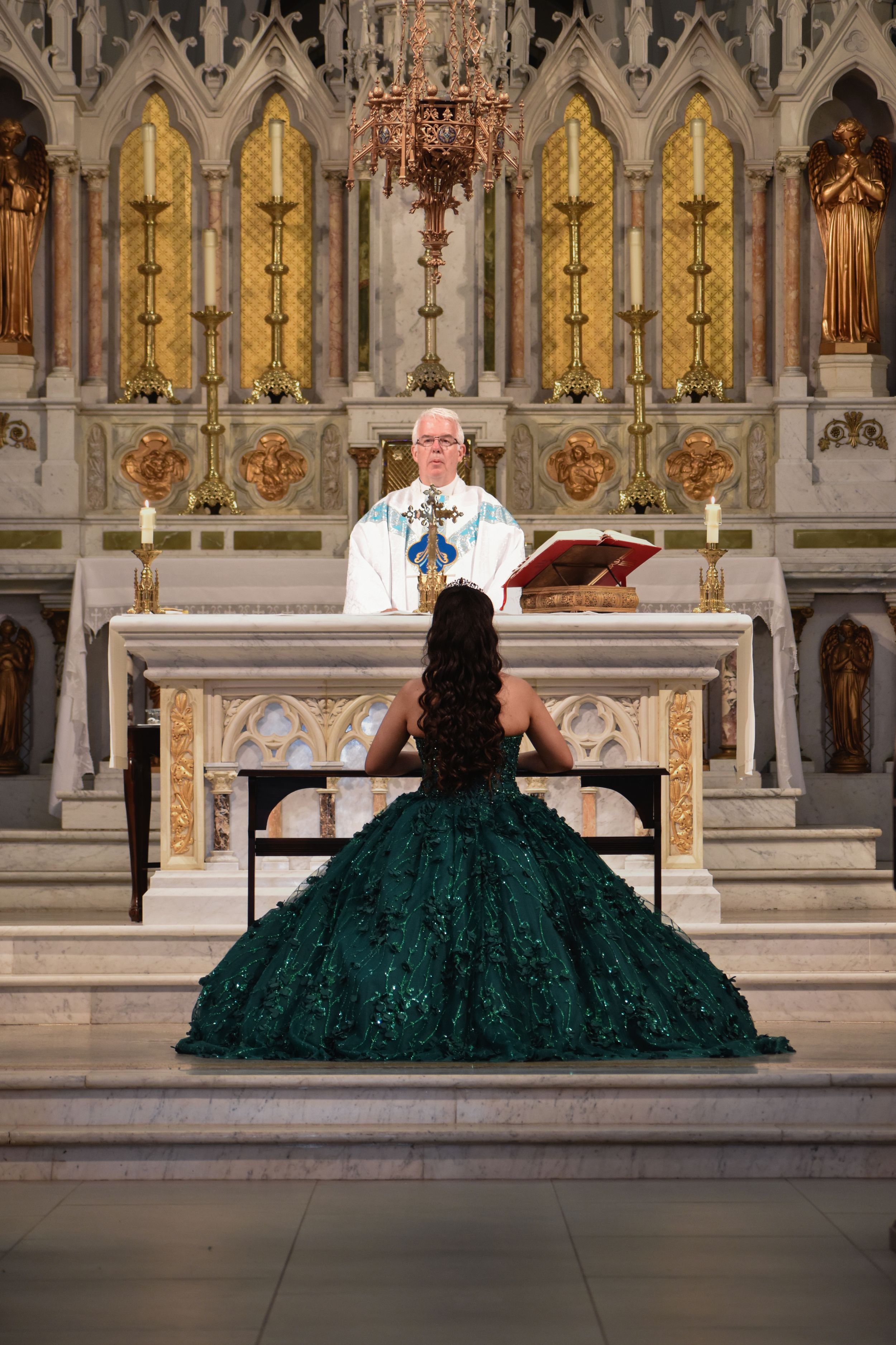 Young woman in emerald green quinceañera gown kneeling before priest at ornate church altar during ceremony. toronto GTA