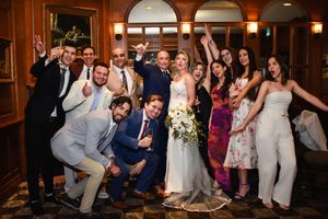 Joyful wedding group photo with bride in white gown and groom in navy suit surrounded by celebrating guests indoors.