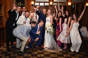 Excited wedding party group celebrating with bride holding white bouquet in elegant wood-paneled room. Toronto