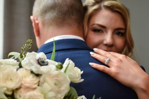 Bride embracing groom in blue suit, holding white floral bouquet with anemones, showcasing diamond engagement ring.