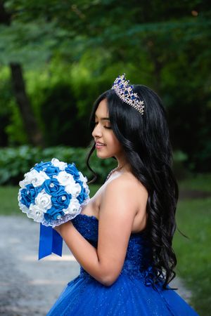 Young Latina girl in royal blue quinceañera gown and tiara holds blue and white rose bouquet outdoors.