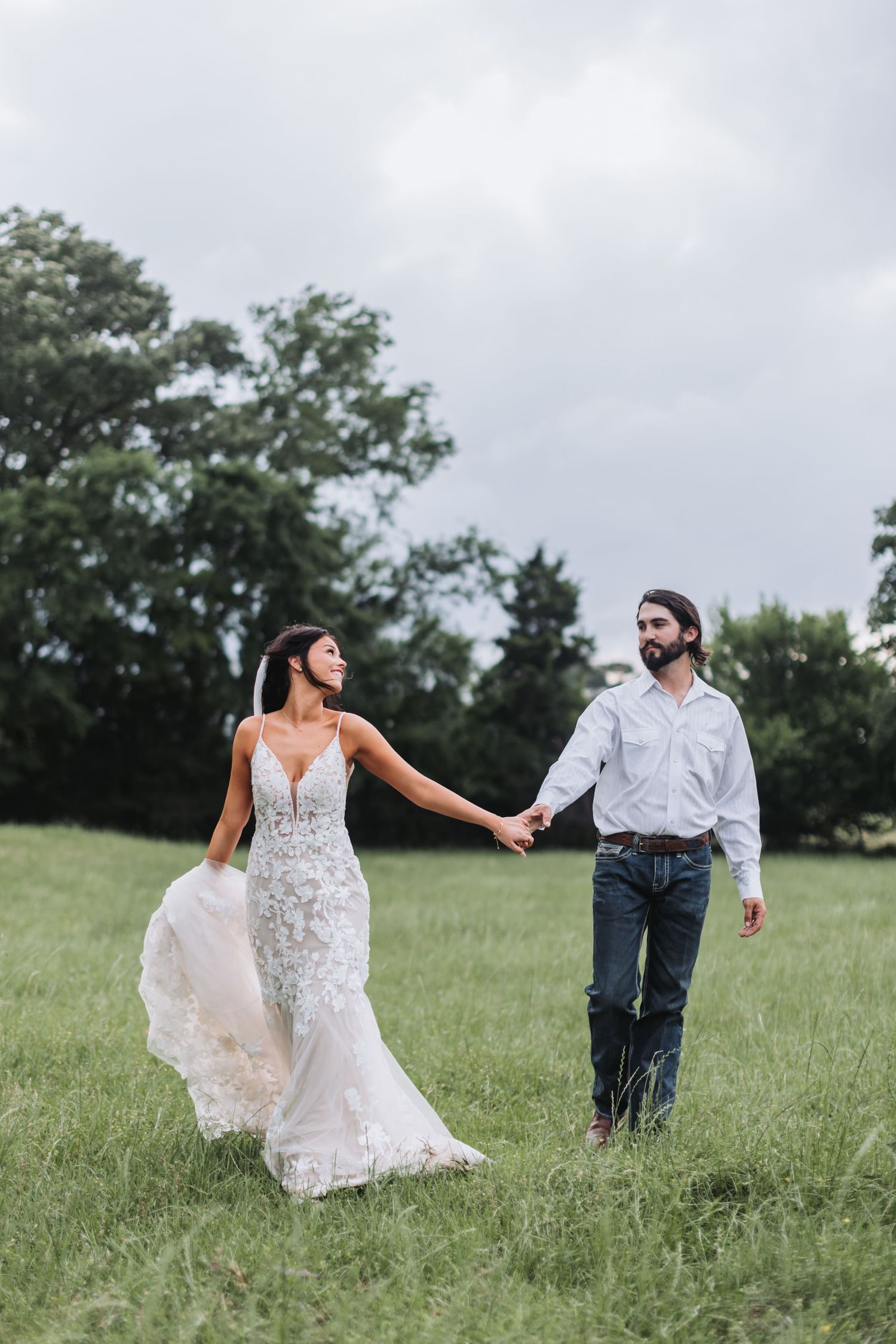 A romantic wedding couple walks hand in hand through a lush green field on an overcast summer day.