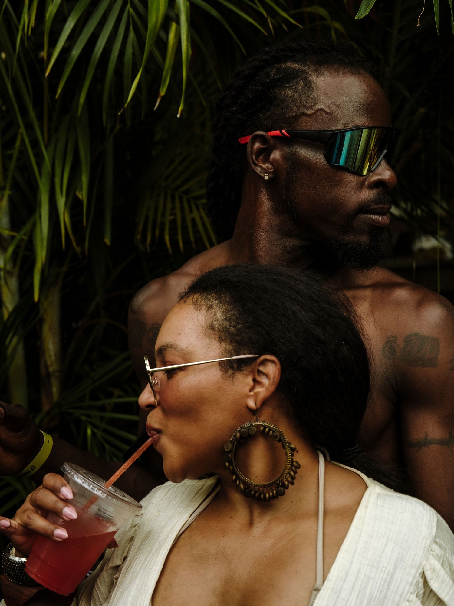 Two people enjoying tropical drinks against a lush palm backdrop wearing stylish sunglasses and jewelry.