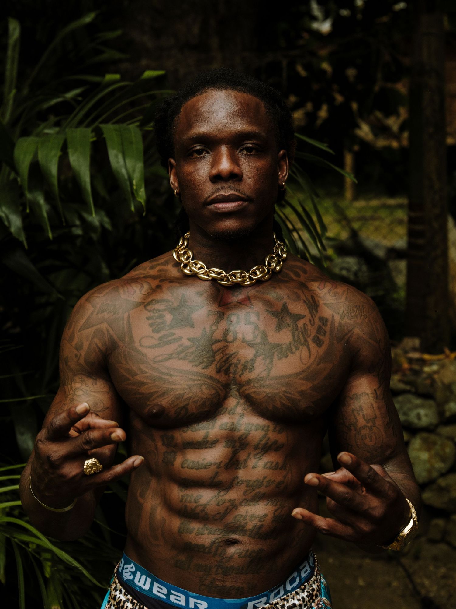 Shirtless muscular person with extensive tattoos and gold chain jewelry poses in dark outdoor setting with foliage background.