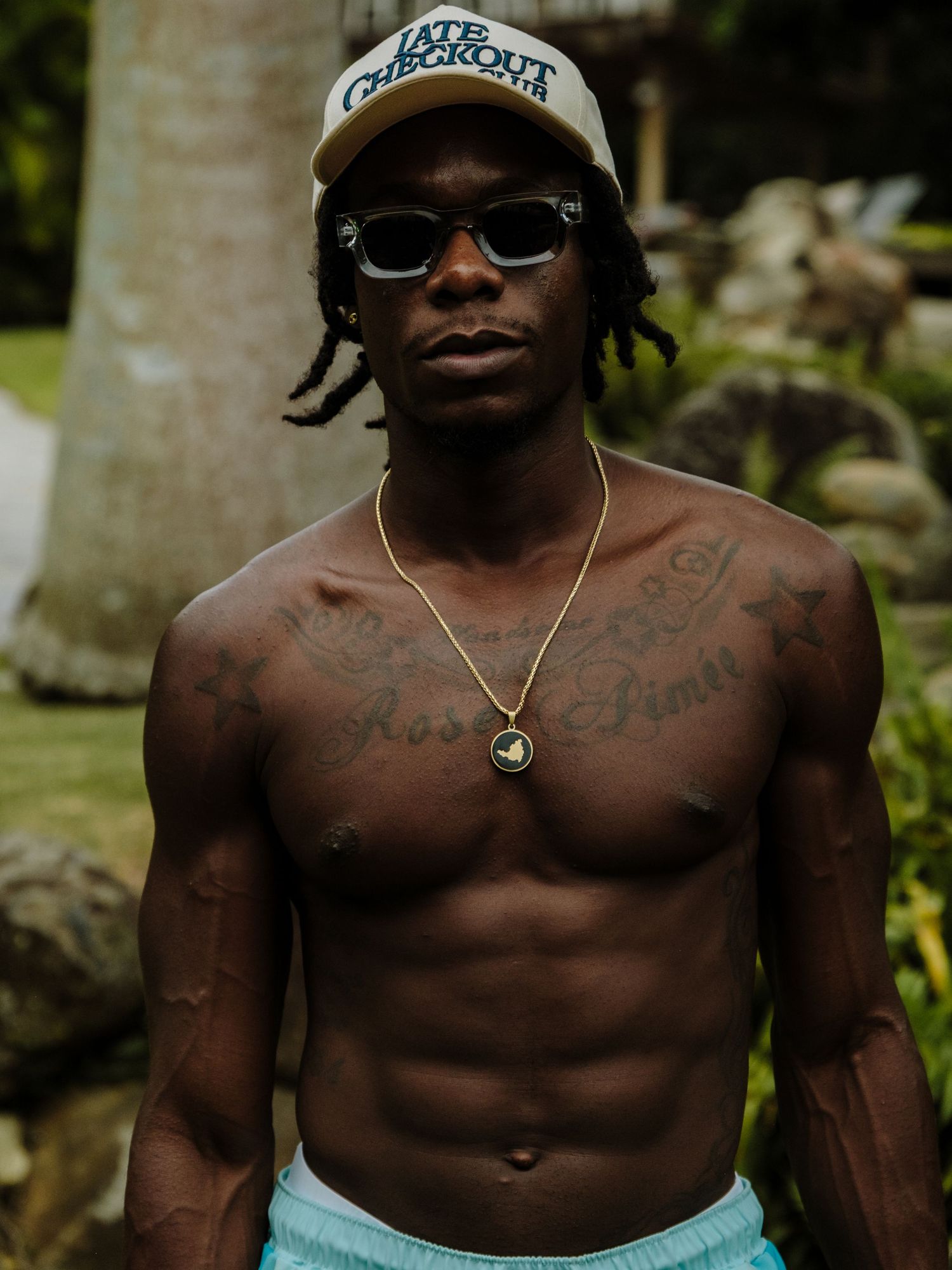 Shirtless person wearing sunglasses and swim trunks poses outside in natural lighting.
