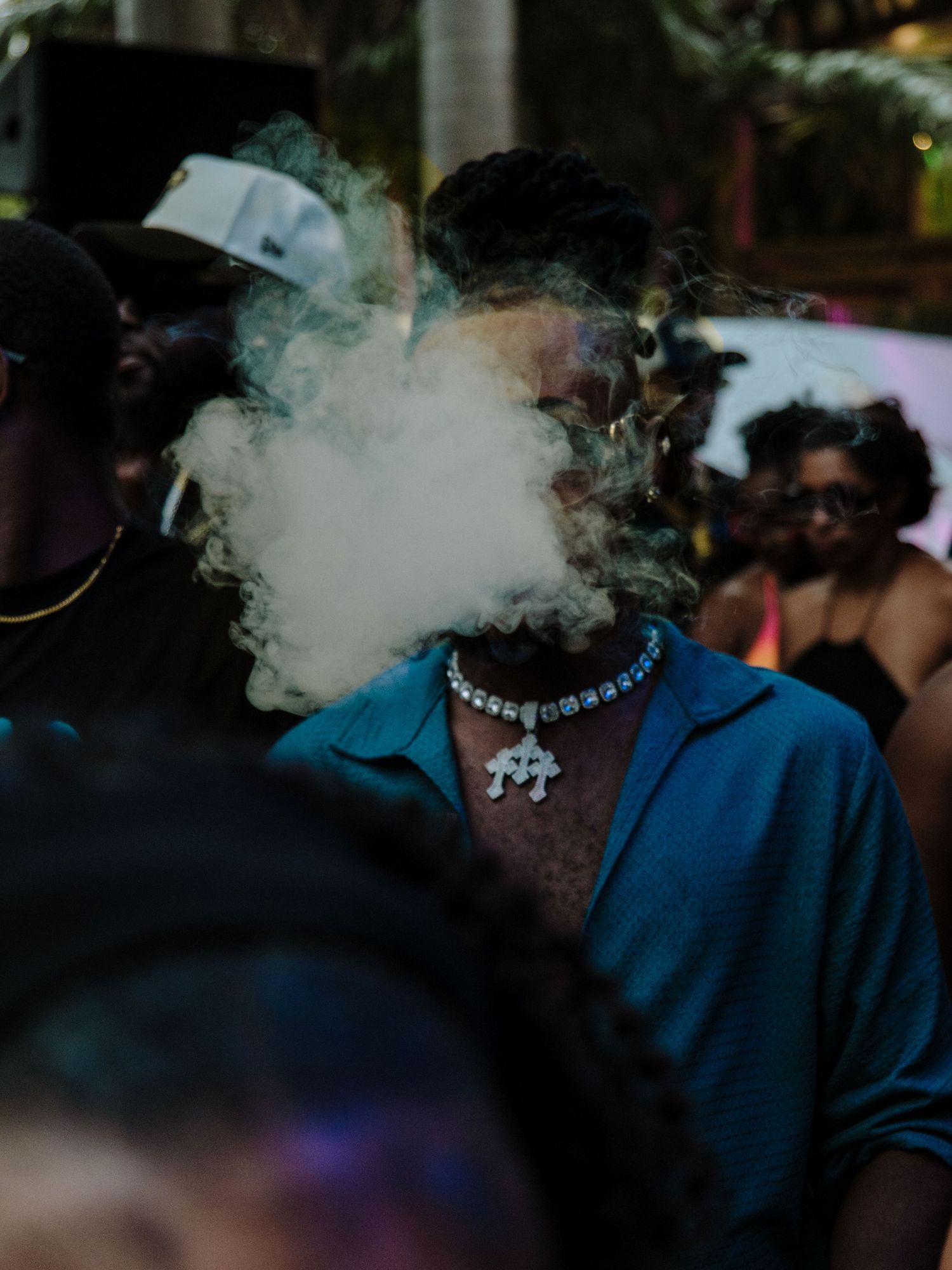 Person wearing blue shirt and necklace exhales smoke in a dimly lit crowd scene.