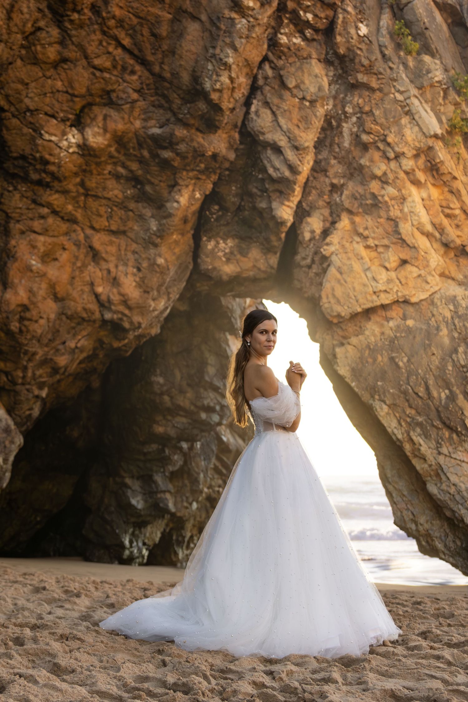 Bride in white ballgown poses under dramatic beach cave arch.