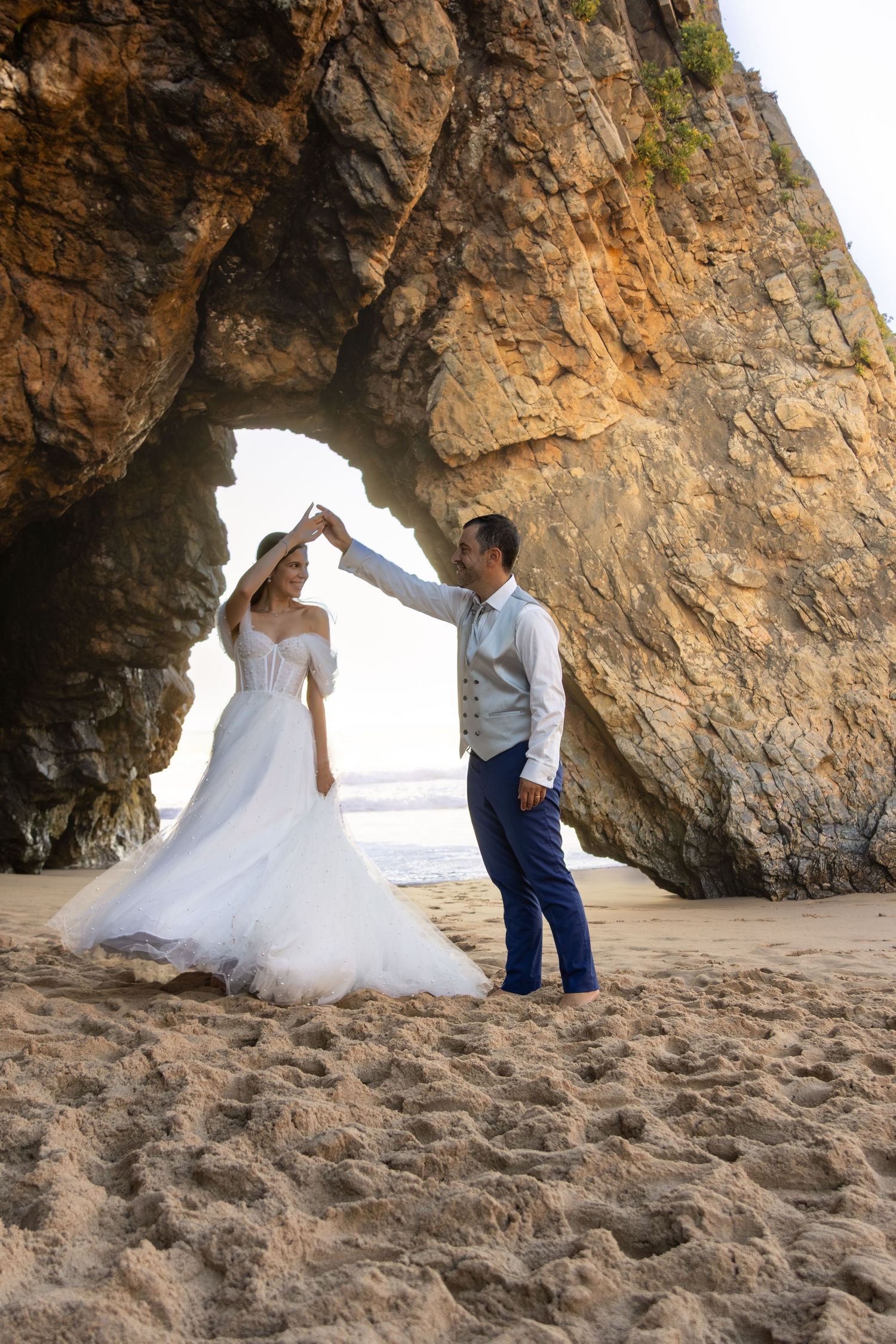 Wedding couple dances beneath a natural rock arch on sandy beach.