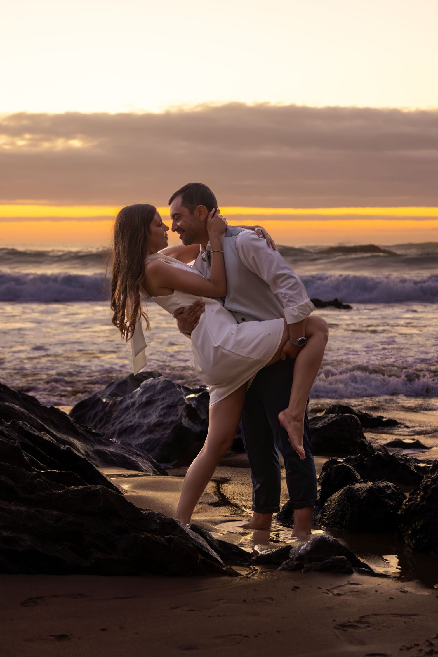 Couple sharing intimate moment on rocky beach during dramatic sunset.