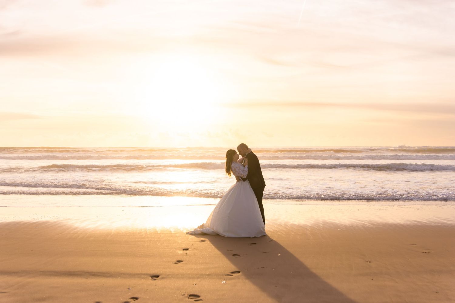 A romantic sunset beach wedding photoshoot with a couple in formal attire silhouetted against the golden sky.