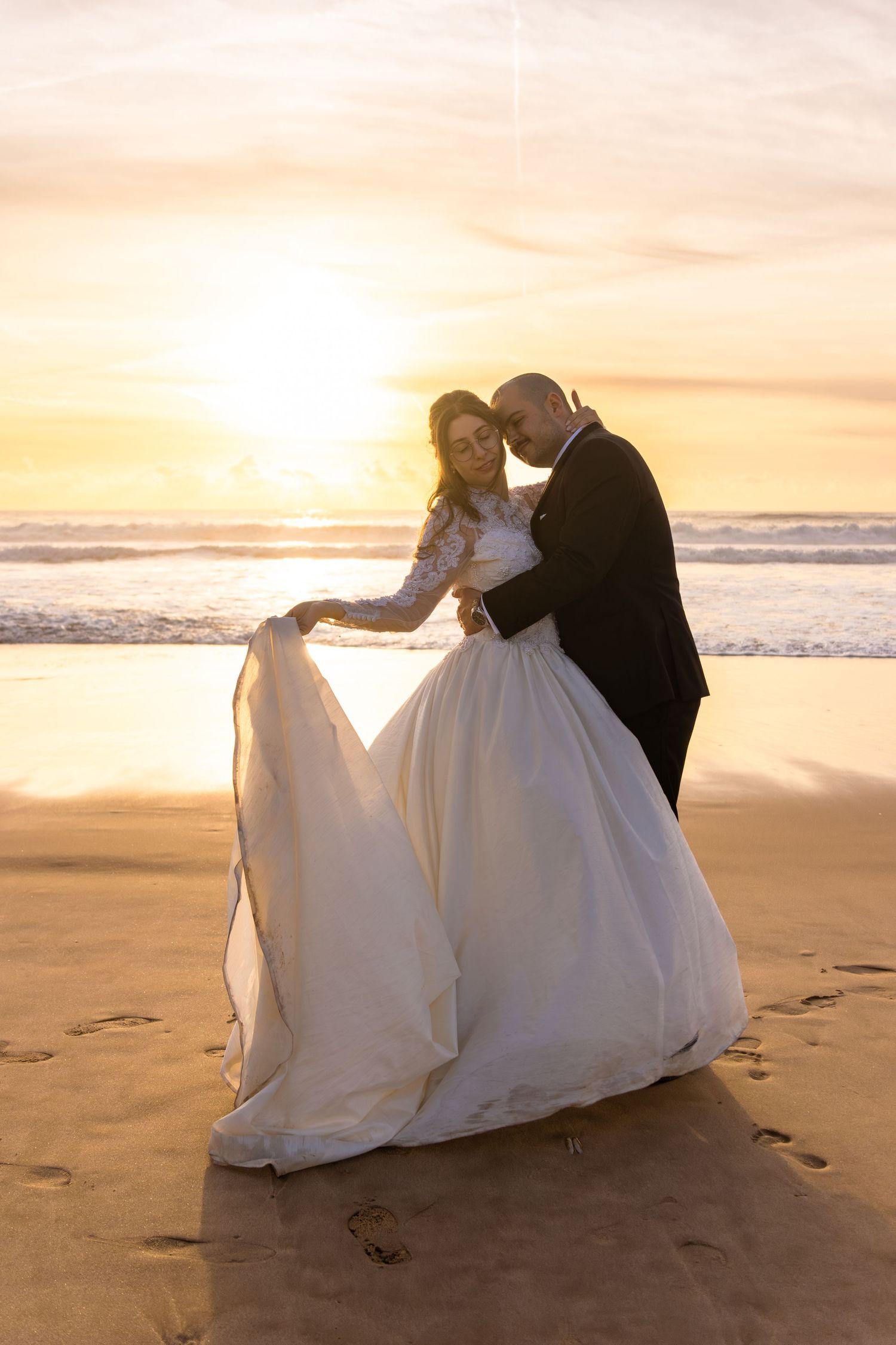 Newlyweds share intimate moments on a sandy beach at sunset with flowing white wedding dress.