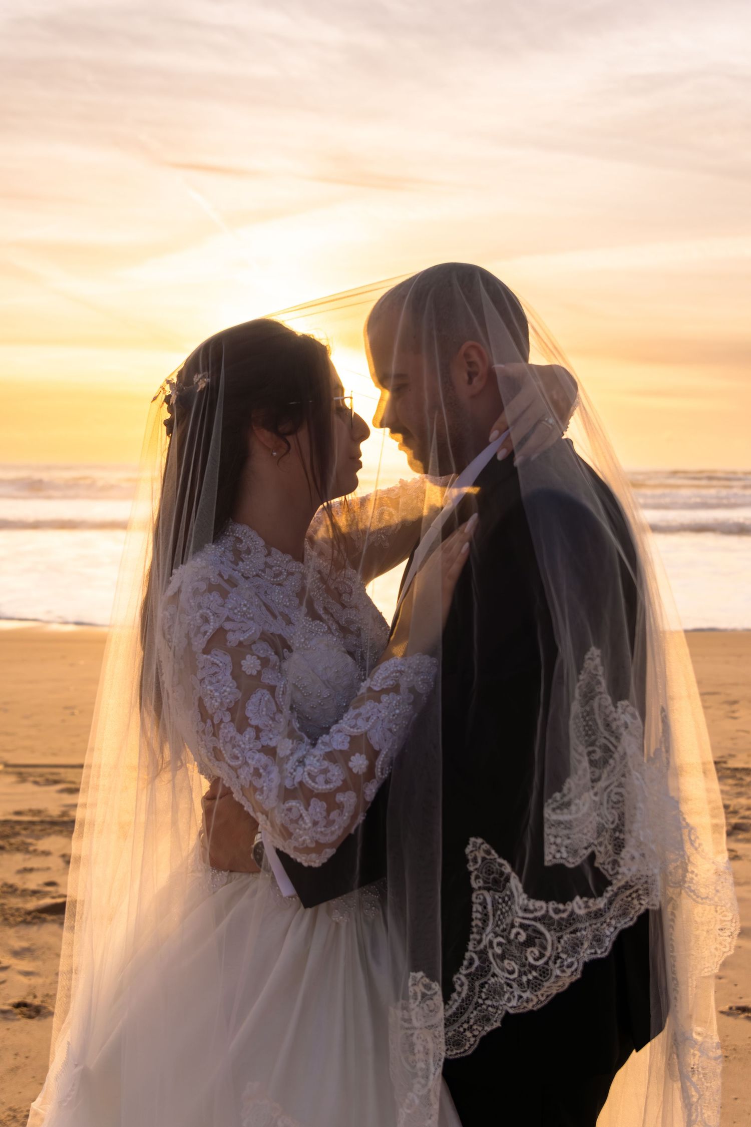 Silhouetted wedding couple shares romantic moments under a flowing veil at sunset on the beach.