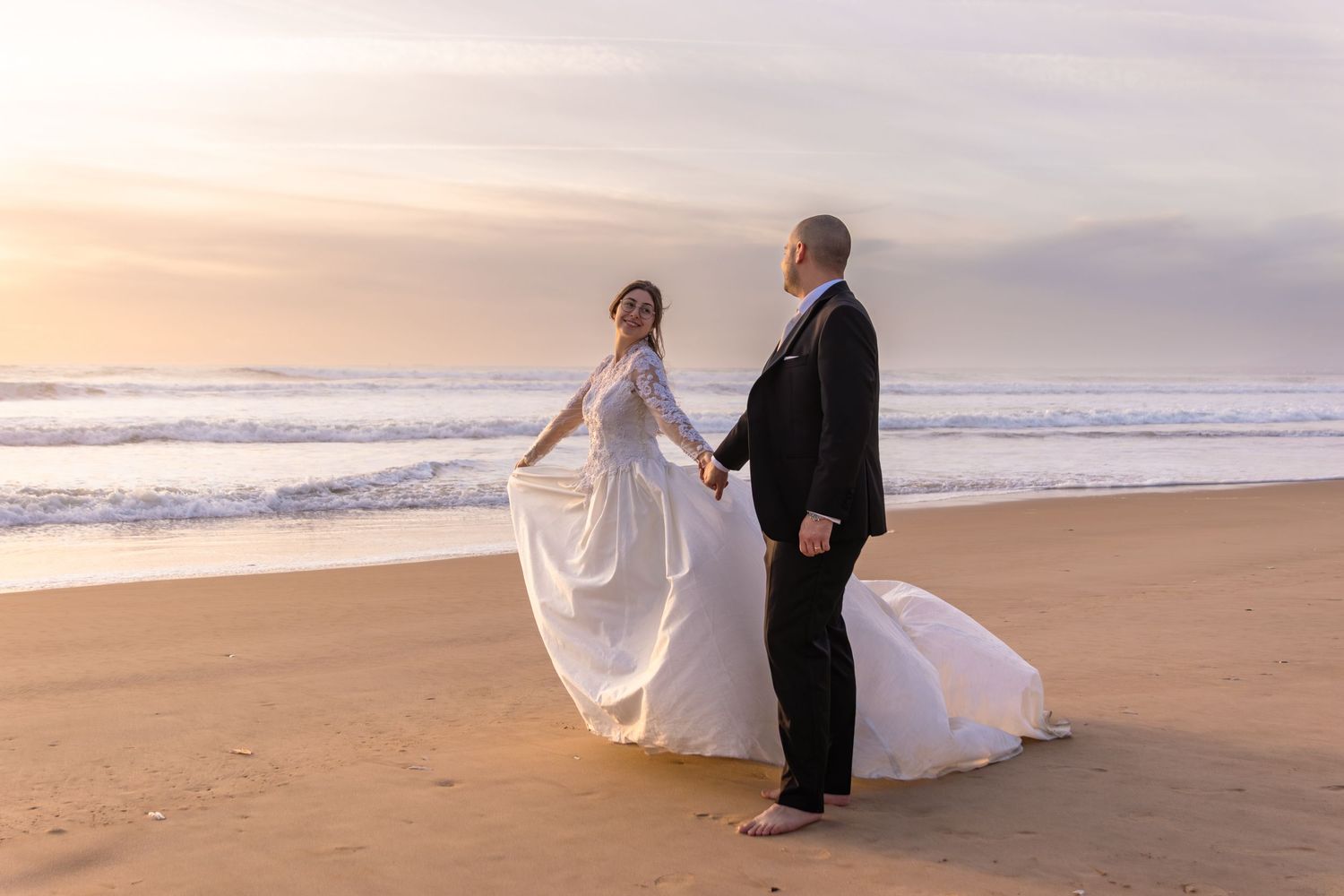 Couple in wedding attire walks along shoreline during sunset beach photoshoot.