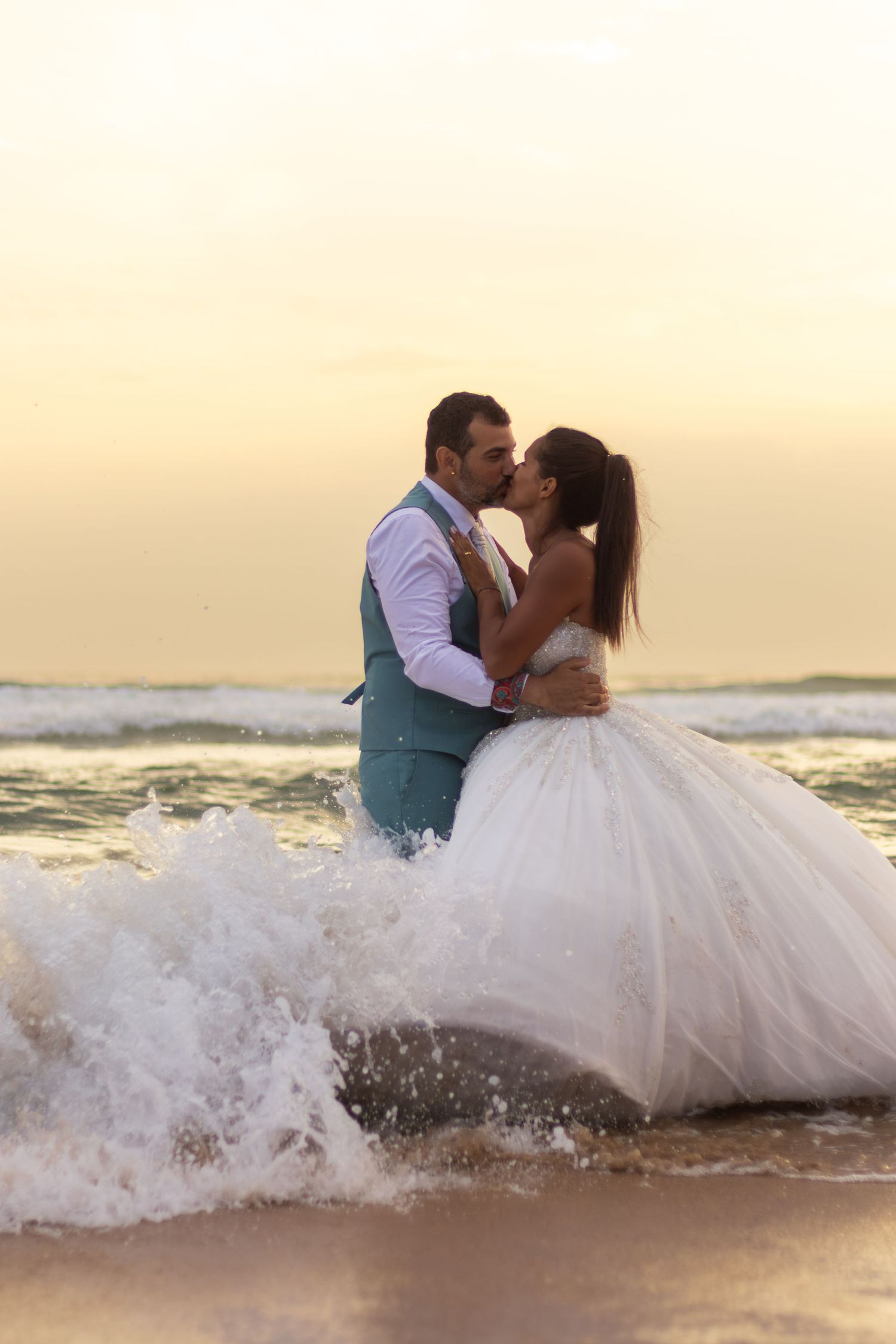Romantic couple embracing in white wedding attire as ocean waves crash around them during sunset.