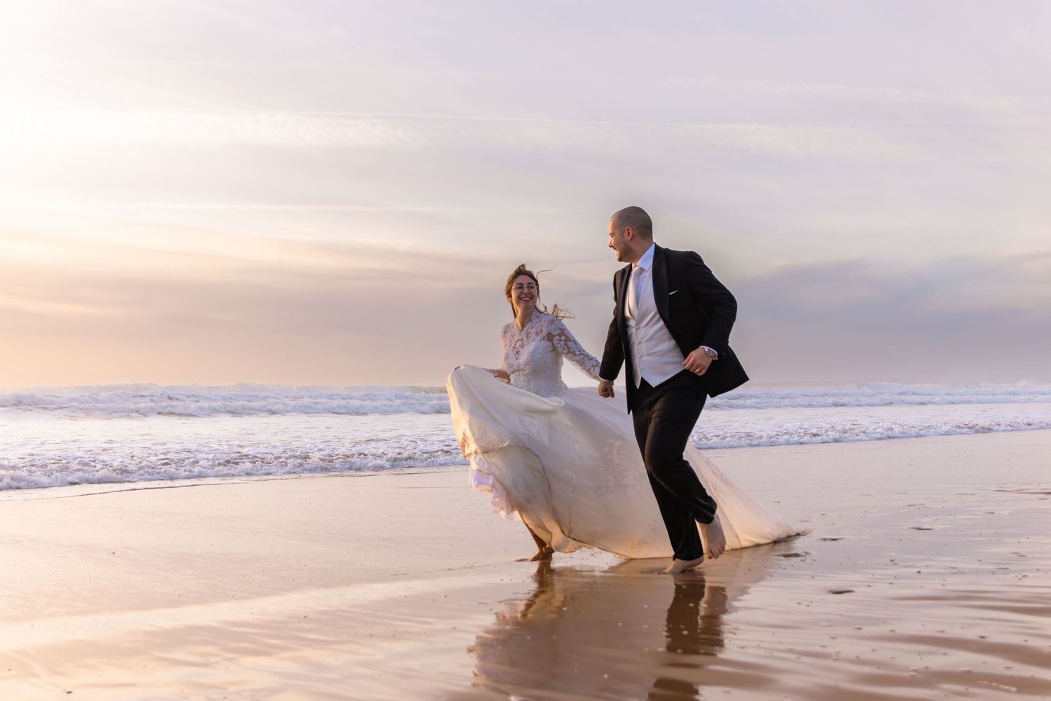Newlyweds dance playfully in the ocean waves at sunset on a beach.