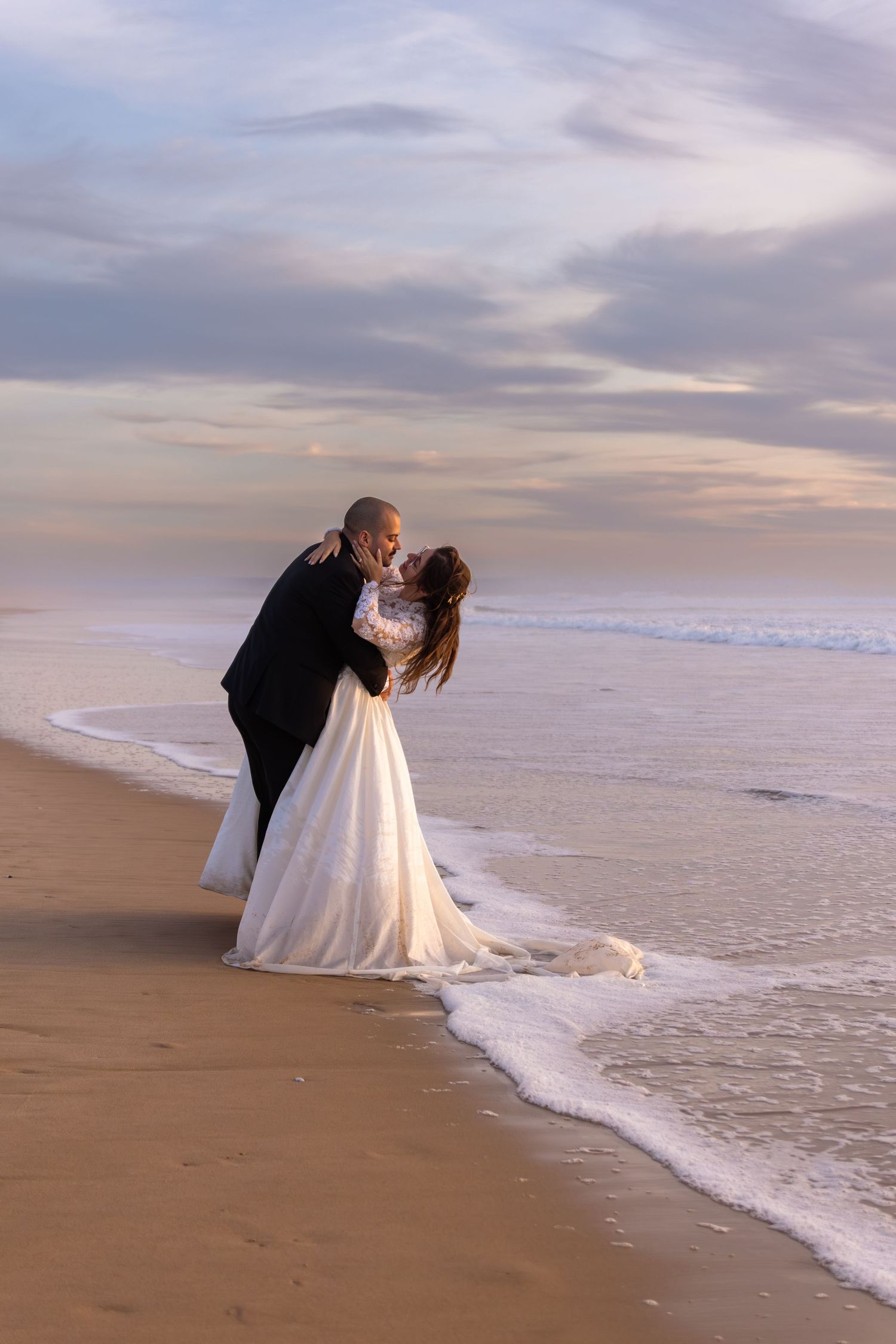 Wedding couple embraces at the edge of the ocean during golden hour.