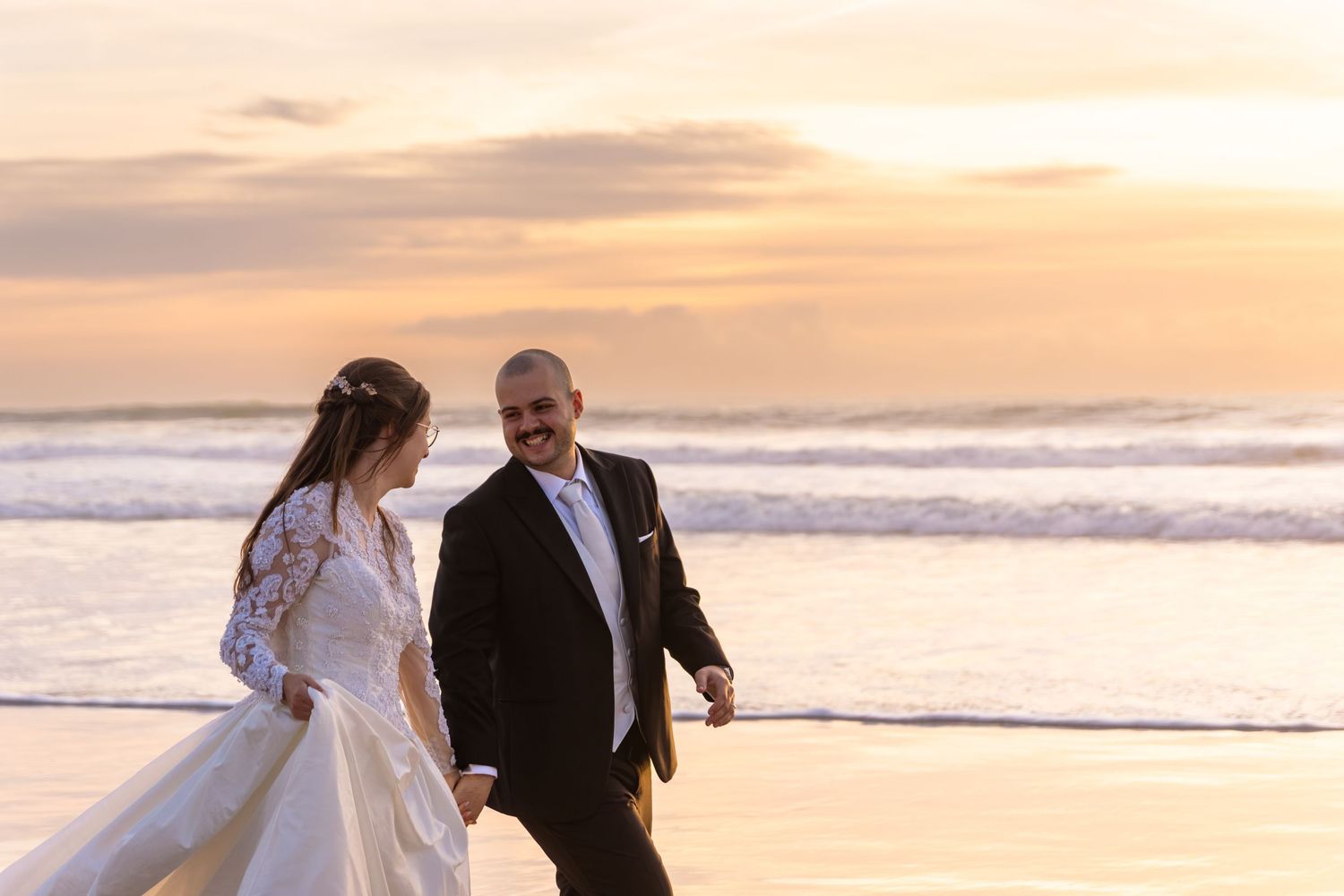 Couple in wedding attire runs joyfully along the beach at sunset.