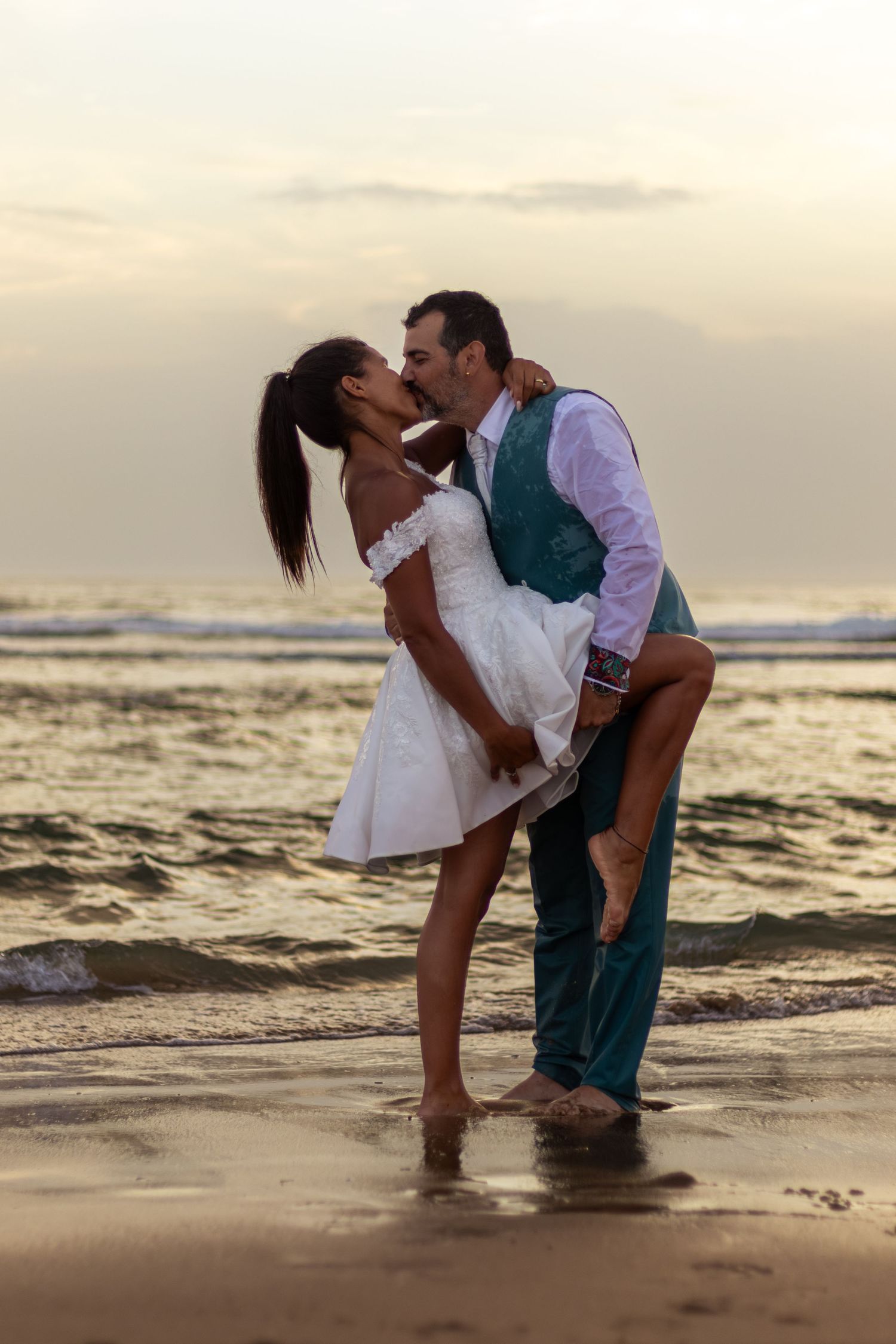 Playful couple in wedding attire dancing in shallow ocean water at dusk.