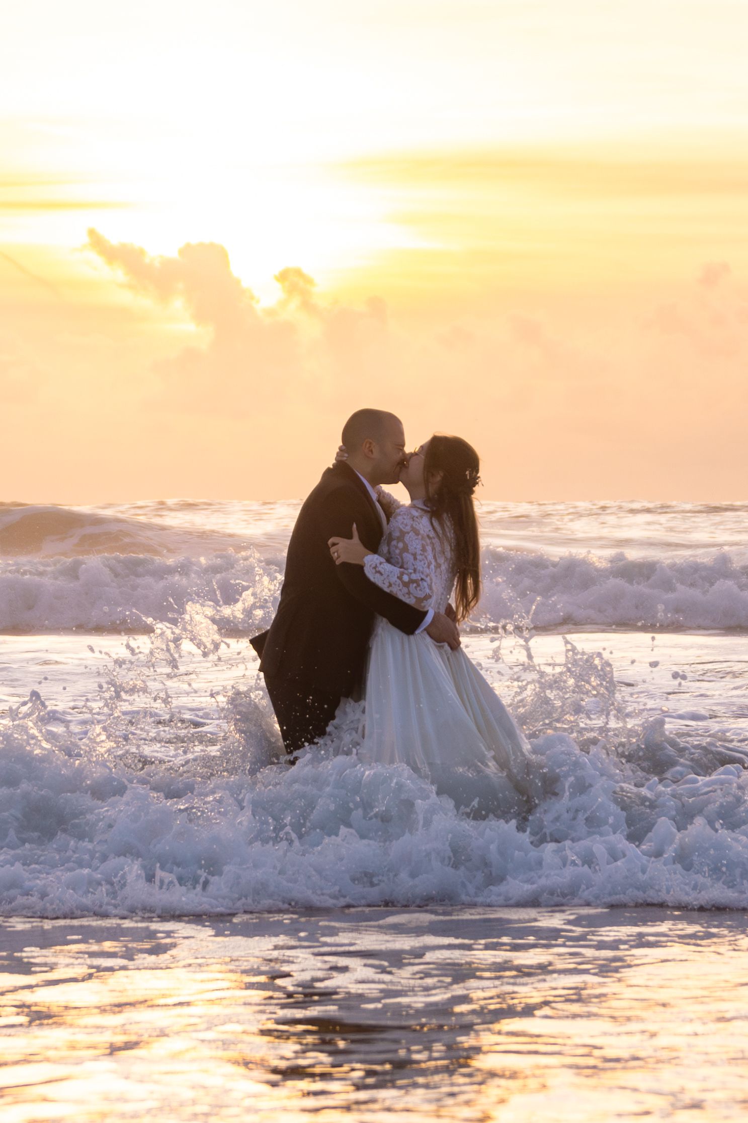 Wedding couple gets splashed by ocean waves during sunset photoshoot.