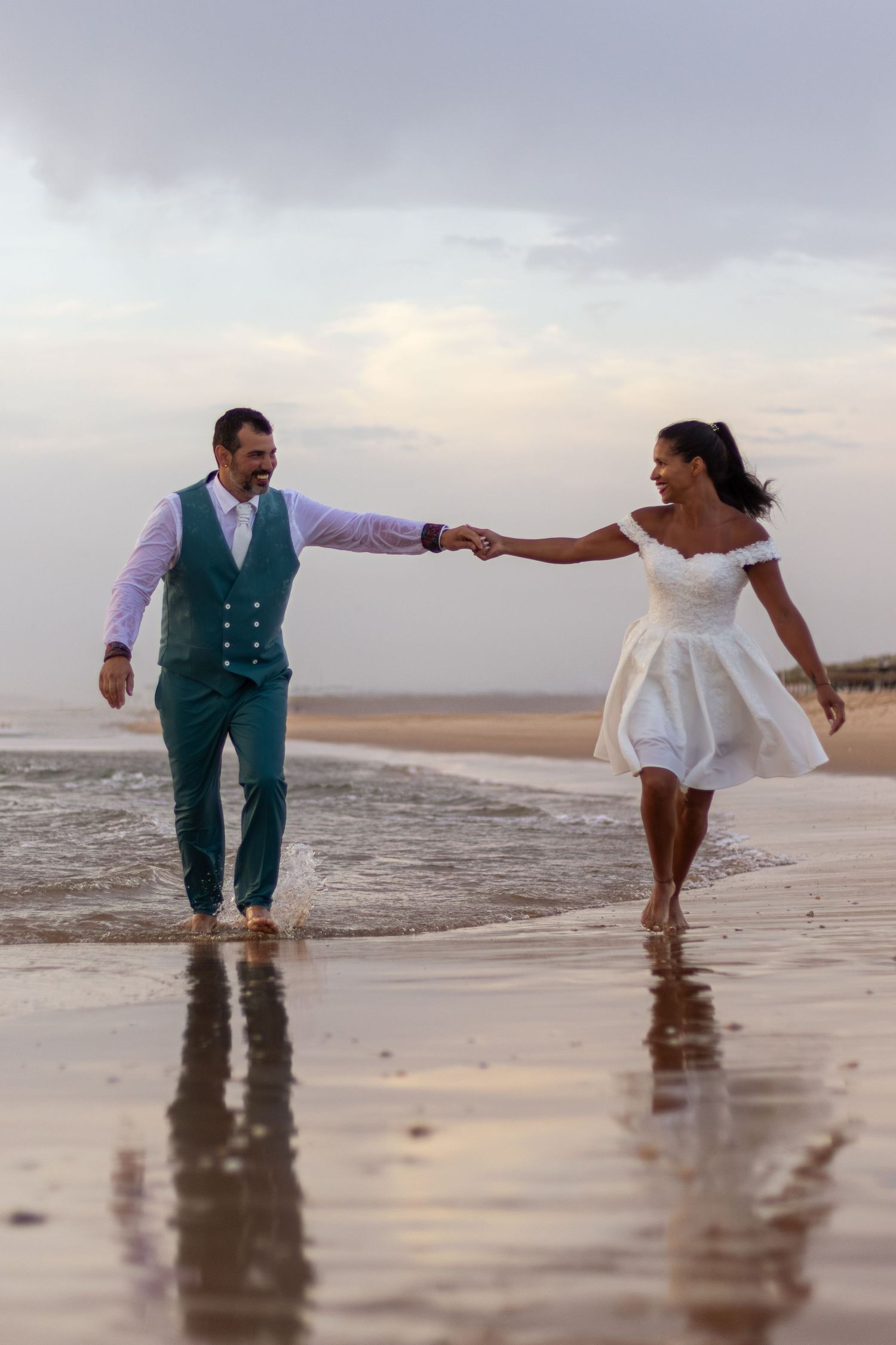 Couple holding hands while walking through shallow beach water at sunset.