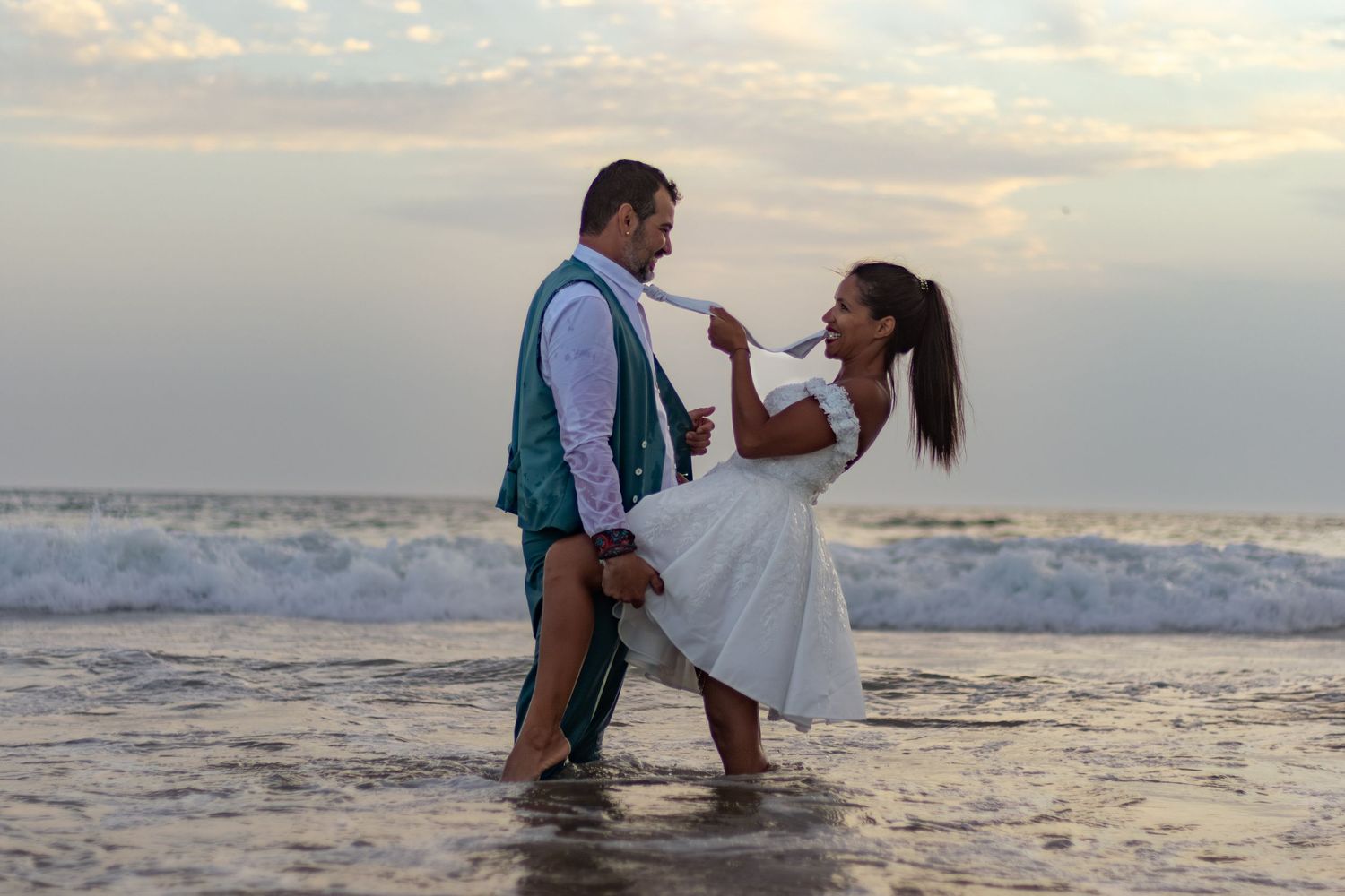 Couple in wedding attire playing in ocean waves during sunset photoshoot.
