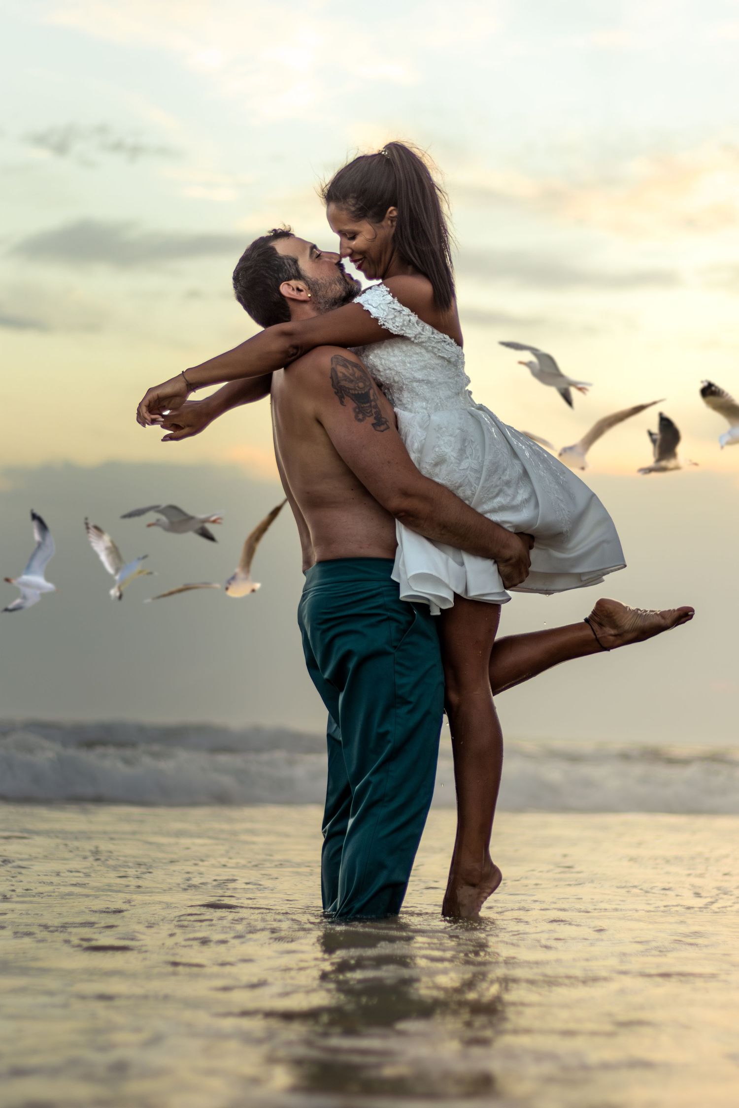 Romantic beach scene with seagulls flying as couple embraces in ocean water.