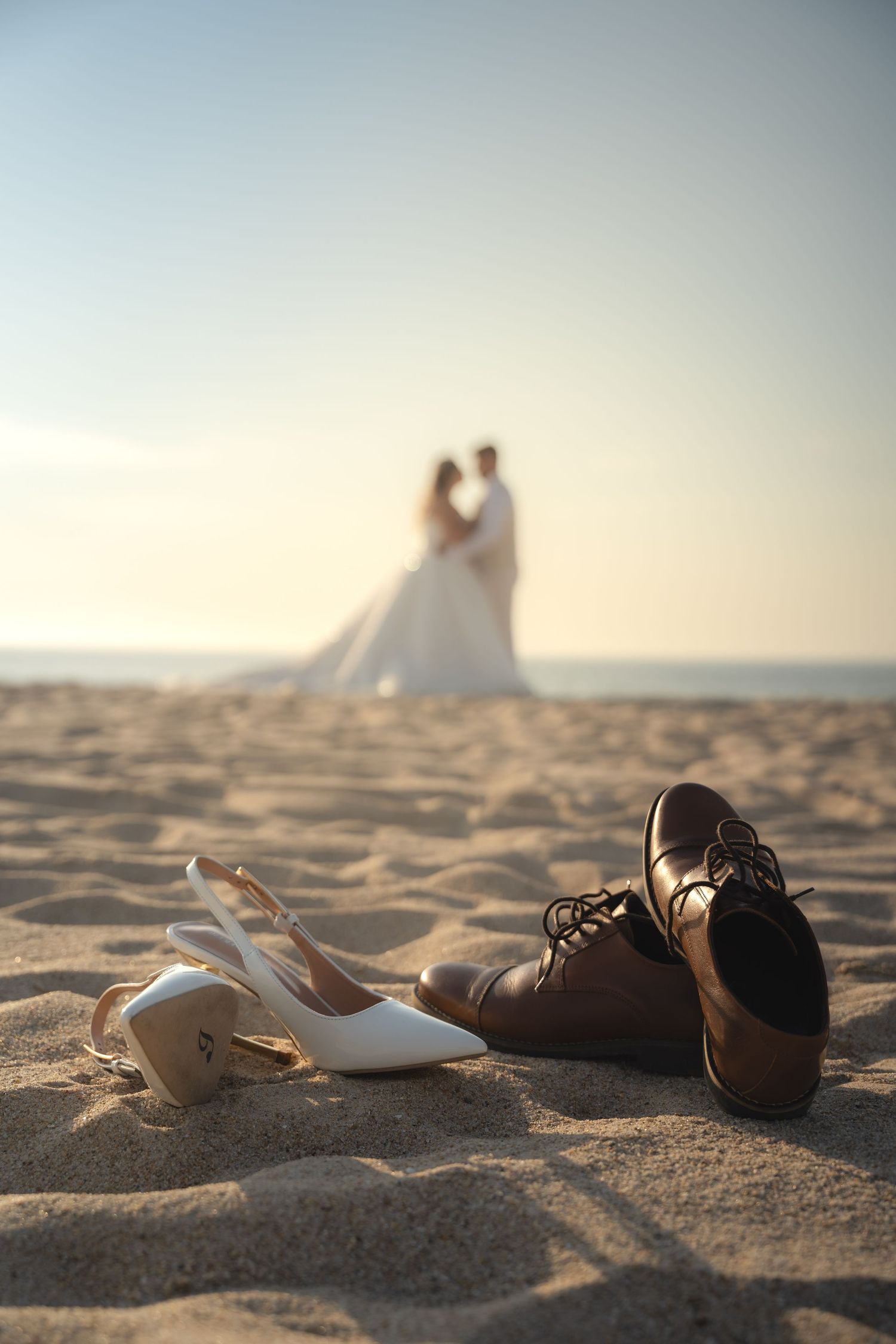 Wedding shoes on sandy beach with blurred newlyweds embracing at sunset in the background.
