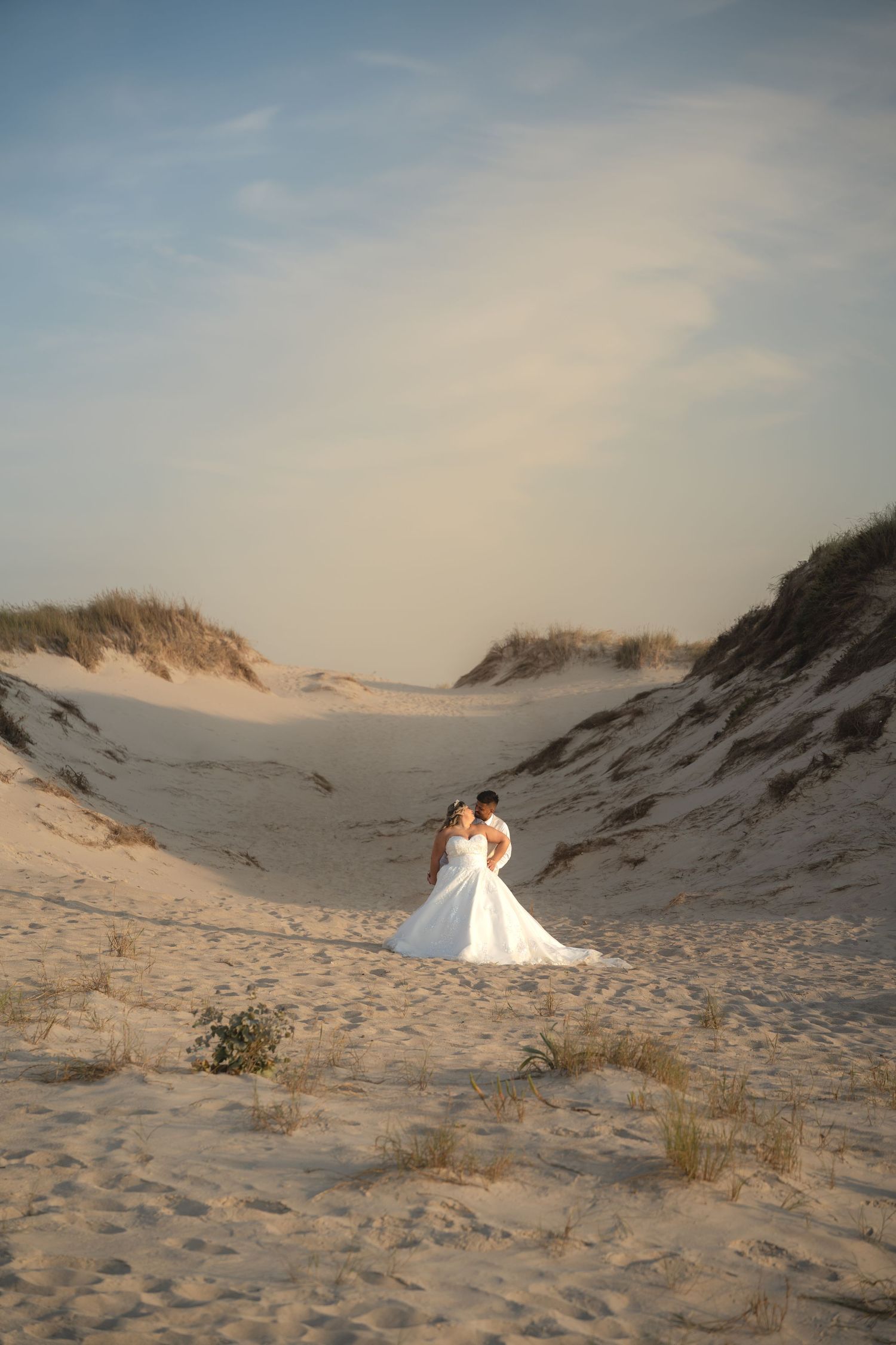 A bride and groom embrace romantically among golden sand dunes at sunset on a beautiful beach wedding day.
