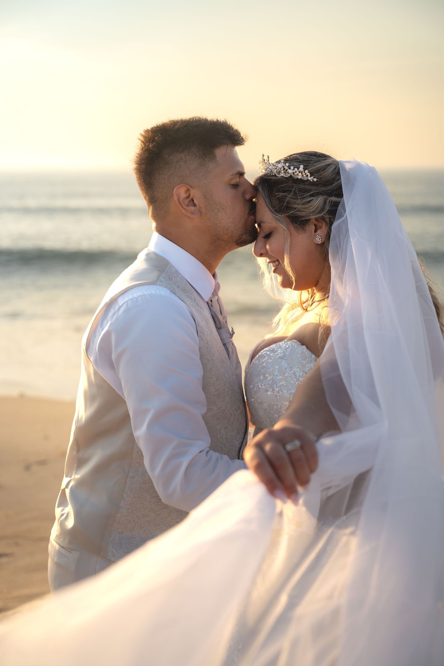 Romantic newlyweds share a tender kiss on the beach at sunset, bride in white gown and veil with tiara.