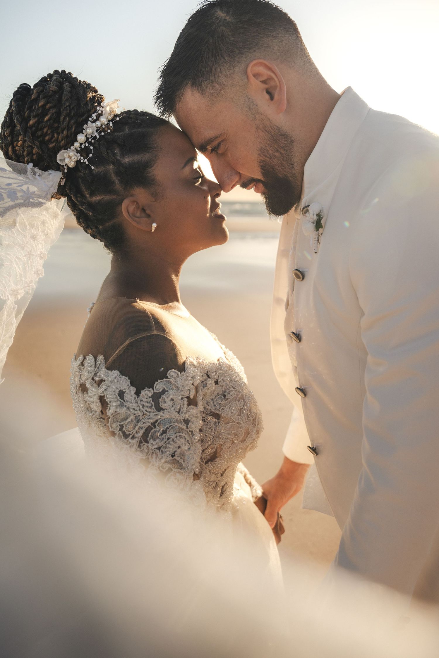 Interracial couple in wedding attire share a romantic moment on a sunlit beach, bride in lace gown and groom in white suit.
