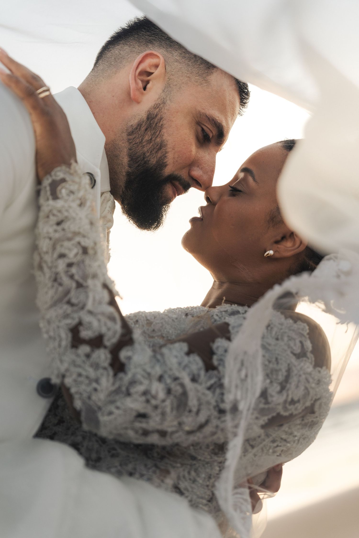 A bride and groom in elegant white wedding attire share a romantic intimate moment, nearly kissing under a flowing veil.