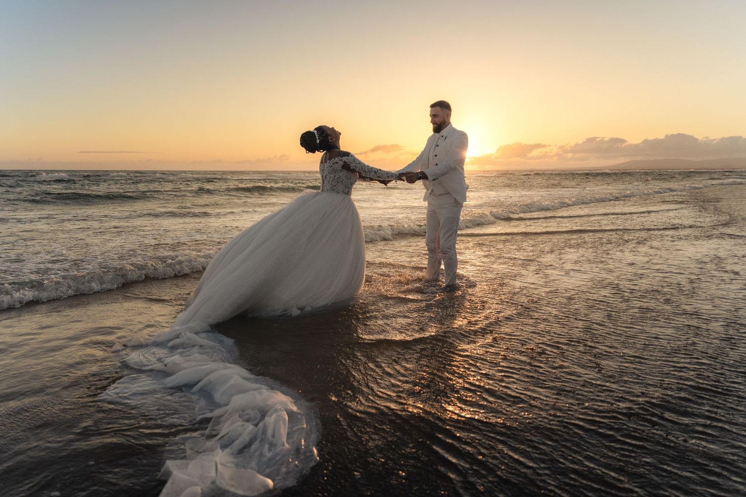 Couple in wedding attire dancing on a beach at sunset with ocean waves and golden sky backdrop.