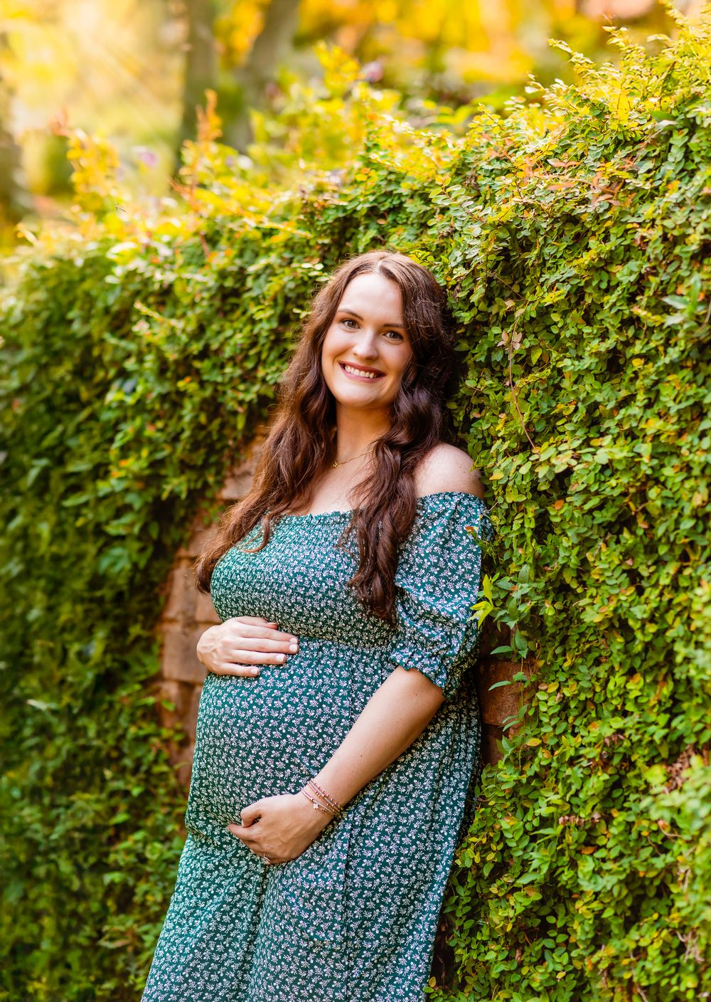 A woman in a blue floral dress stands in front of a lush green hedge wall in outdoor natural lighting.