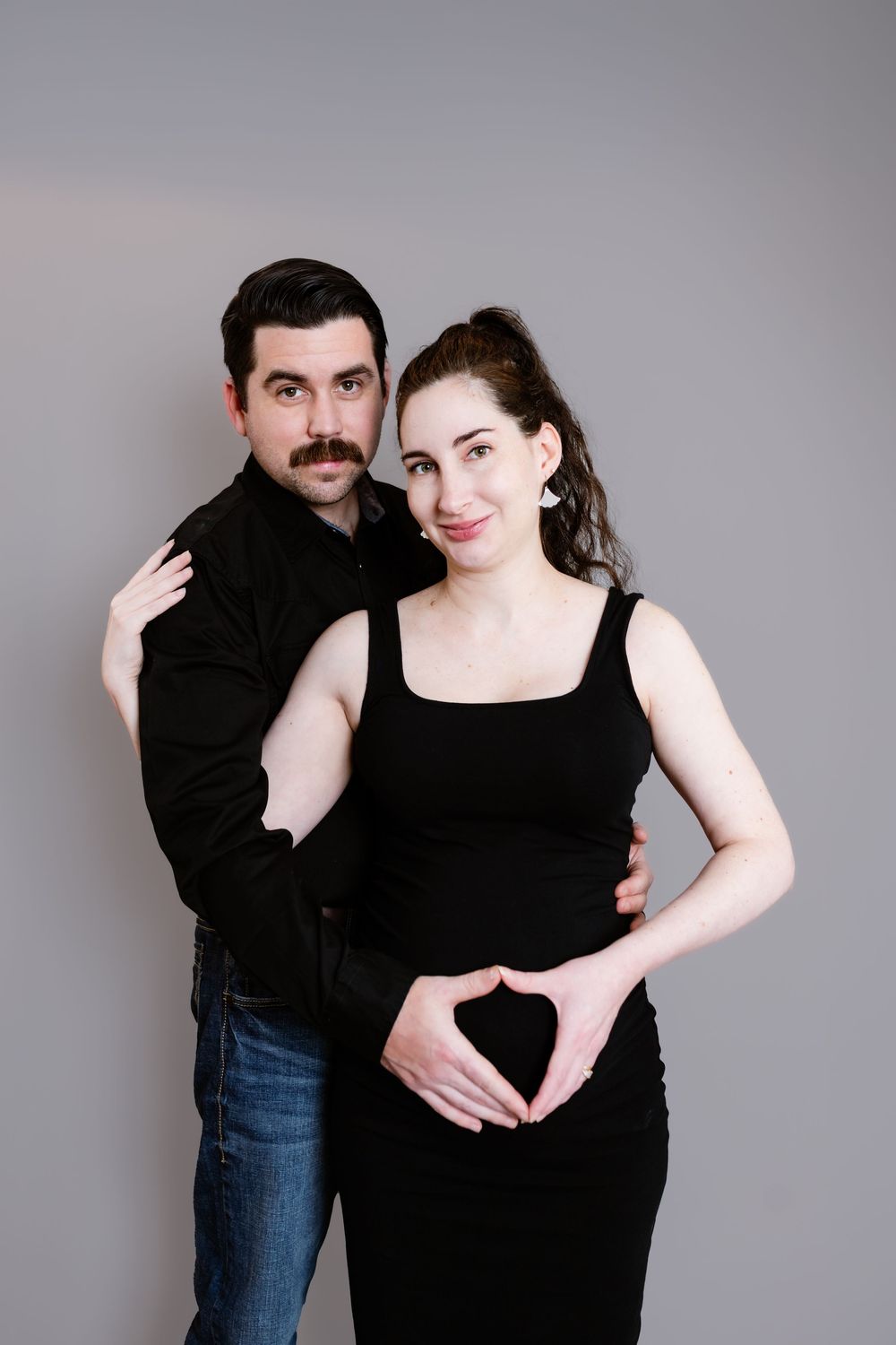 A couple in black attire poses together making a heart shape with their hands against a gray background.