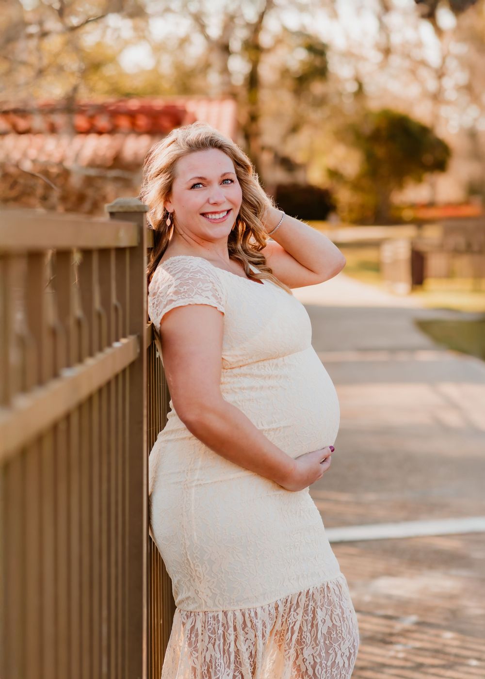 Pregnant woman in white lace dress poses outdoors during golden hour autumn photoshoot.