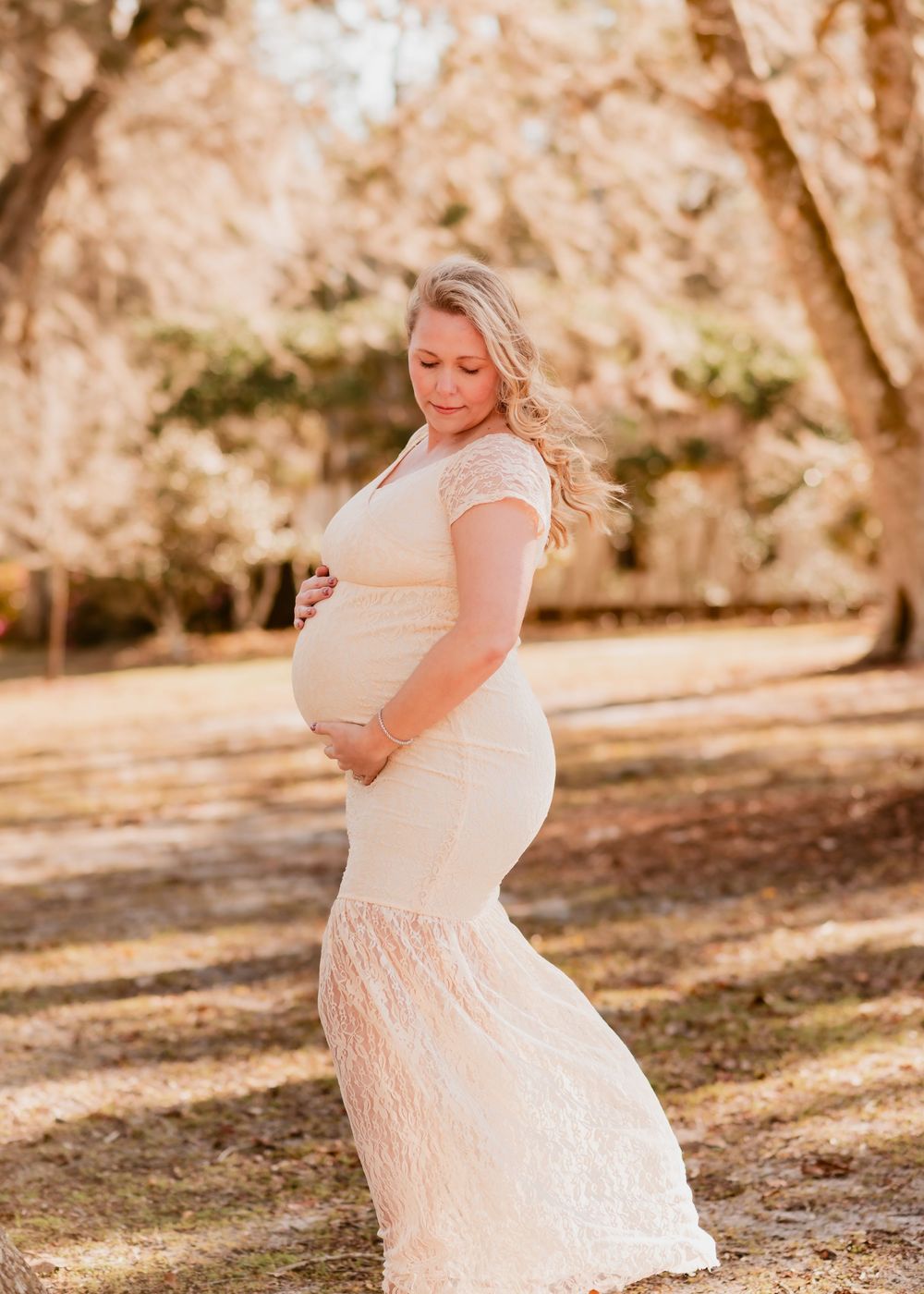 A pregnant woman in a white lace dress poses in a sunlit outdoor setting with trees in the background.