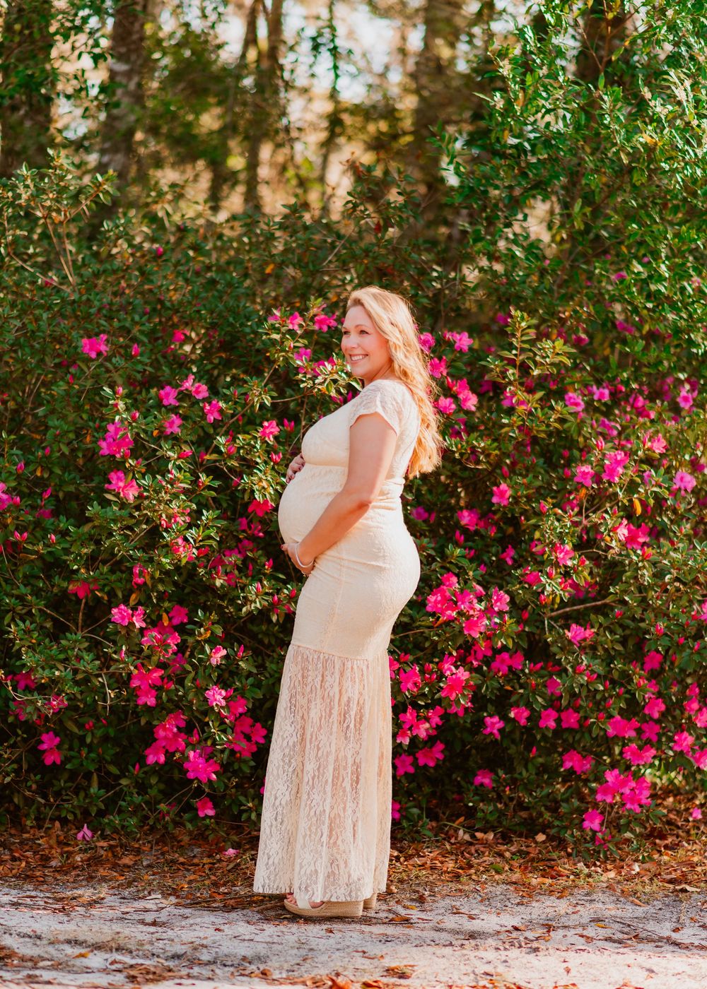 A person in a white lace dress poses against a backdrop of vibrant pink flowers and greenery in a garden setting.
