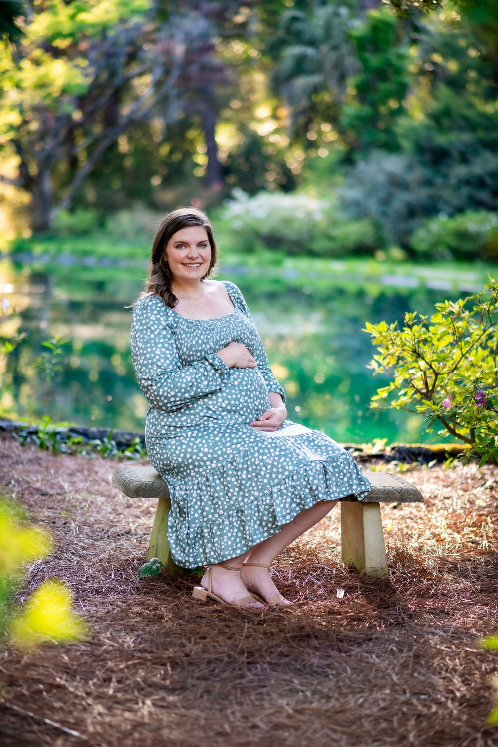 Expectant mother in long patterned dress poses on rustic bench amidst lush garden greenery.
