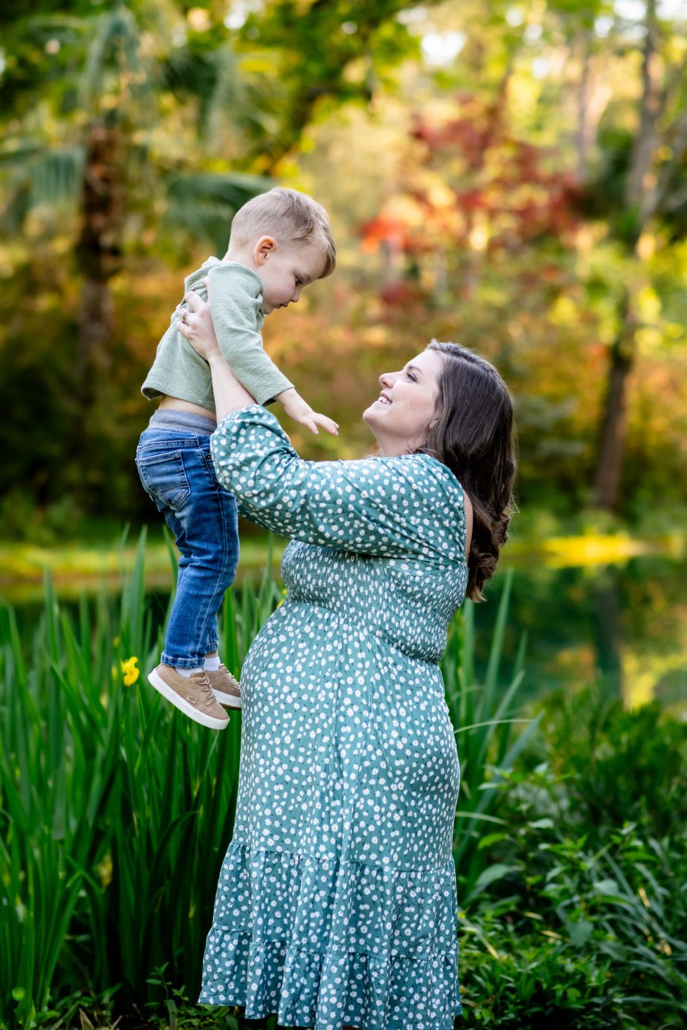Woman in floral dress lifts small child in garden setting with yellow flowers.