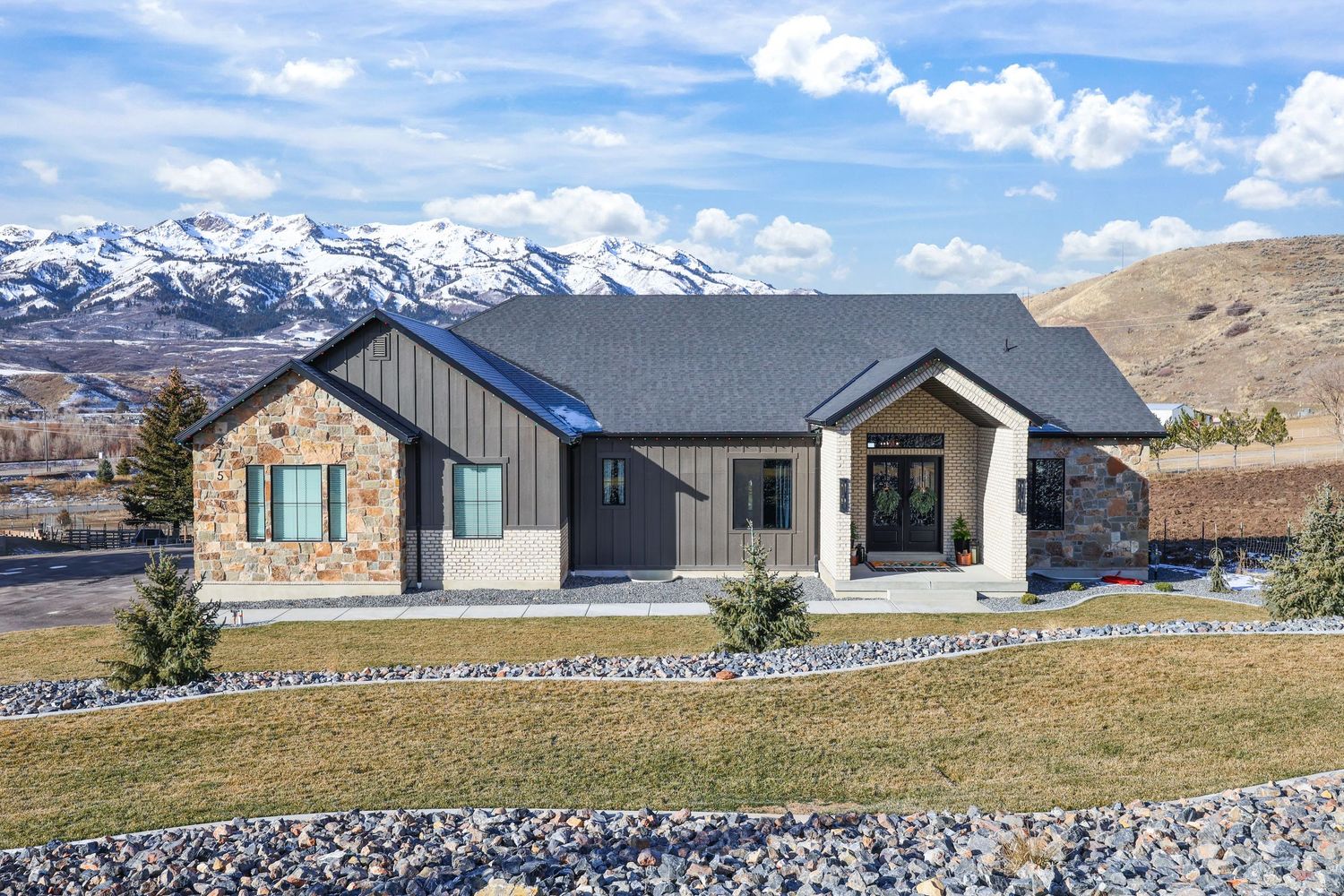 Ranch-style home with stone and board-and-batten exterior, snow-capped mountain backdrop, and lawn. Captured by Slate Media (best in UT)