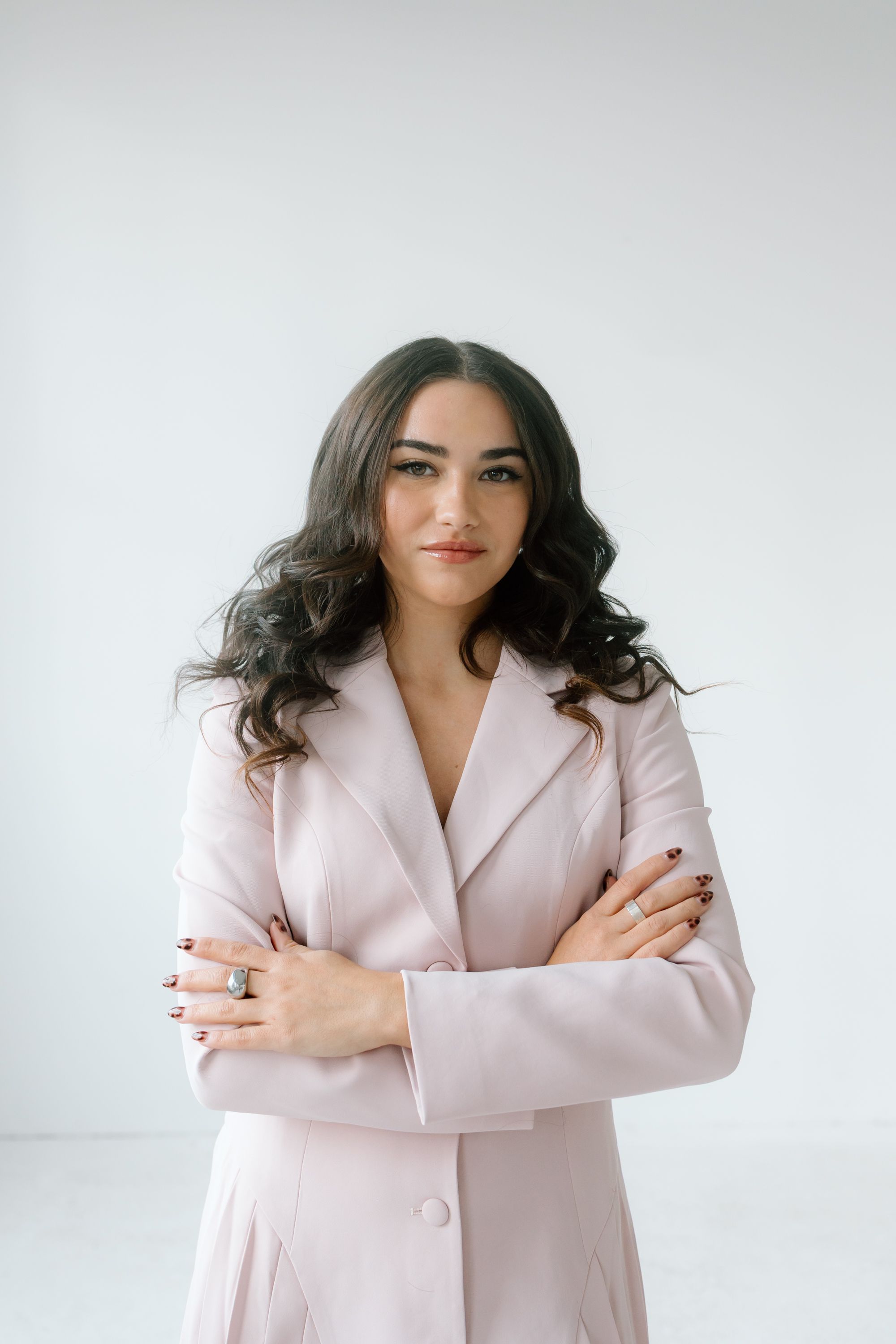 Young woman in pink blazer with crossed arms, dark wavy hair, posing confidently against white background.