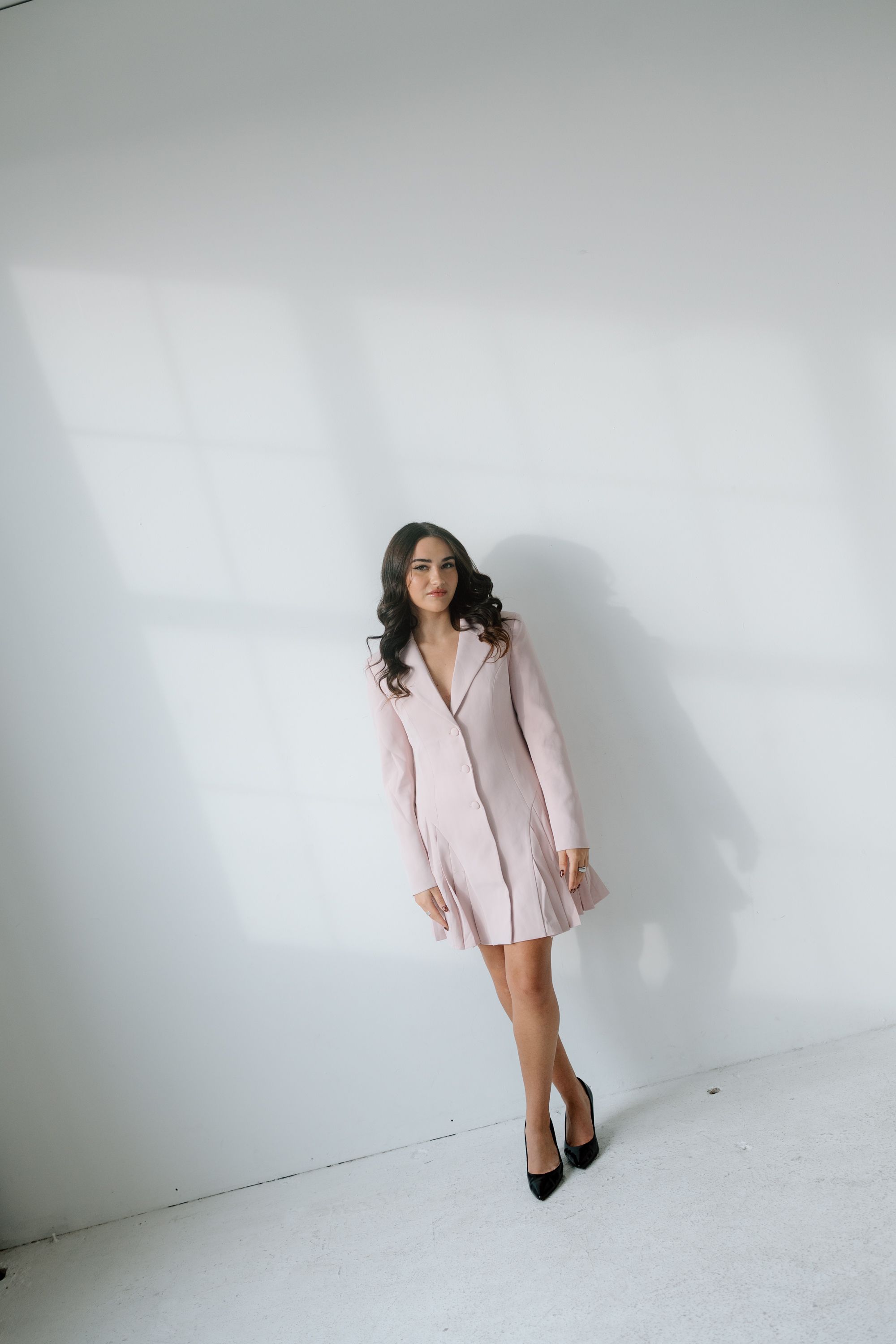 Woman in pink blazer dress and black heels posing against white wall.