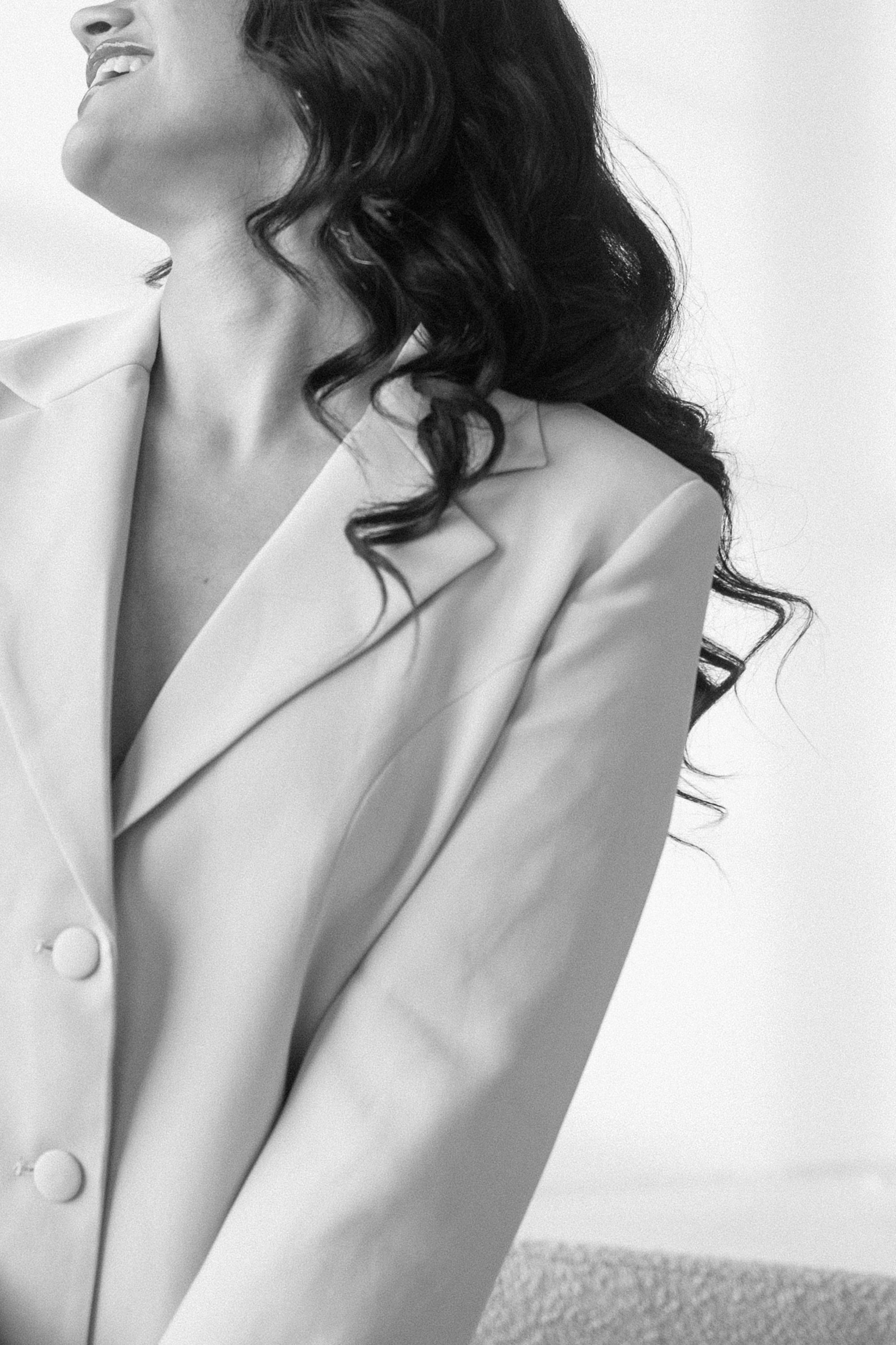 Black and white photo of a woman in a white blazer with dark curly hair, smiling elegantly.