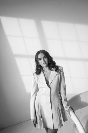 Black and white photo of a young woman in an elegant blazer dress posing in a sunlit studio.