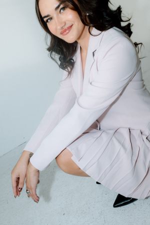 Young woman in light pink blazer and skirt crouching gracefully, smiling at camera in bright studio setting.