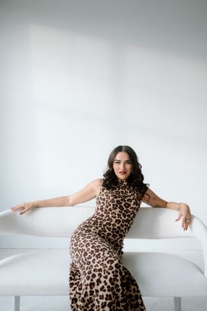 Brunette woman in leopard print dress posing on white sofa in bright minimalist room.