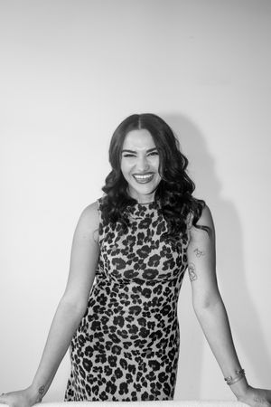 Black and white photo of smiling brunette woman in leopard print dress leaning on white surface.