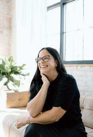 Relaxed indoor portrait showing someone in black attire seated on beige couch near window.